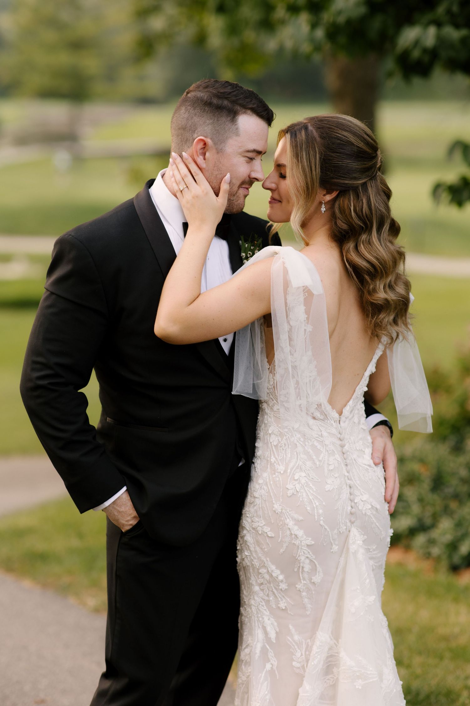 Bride and groom share a romantic moment outdoors, with bride in lace backless gown and groom in black tuxedo.