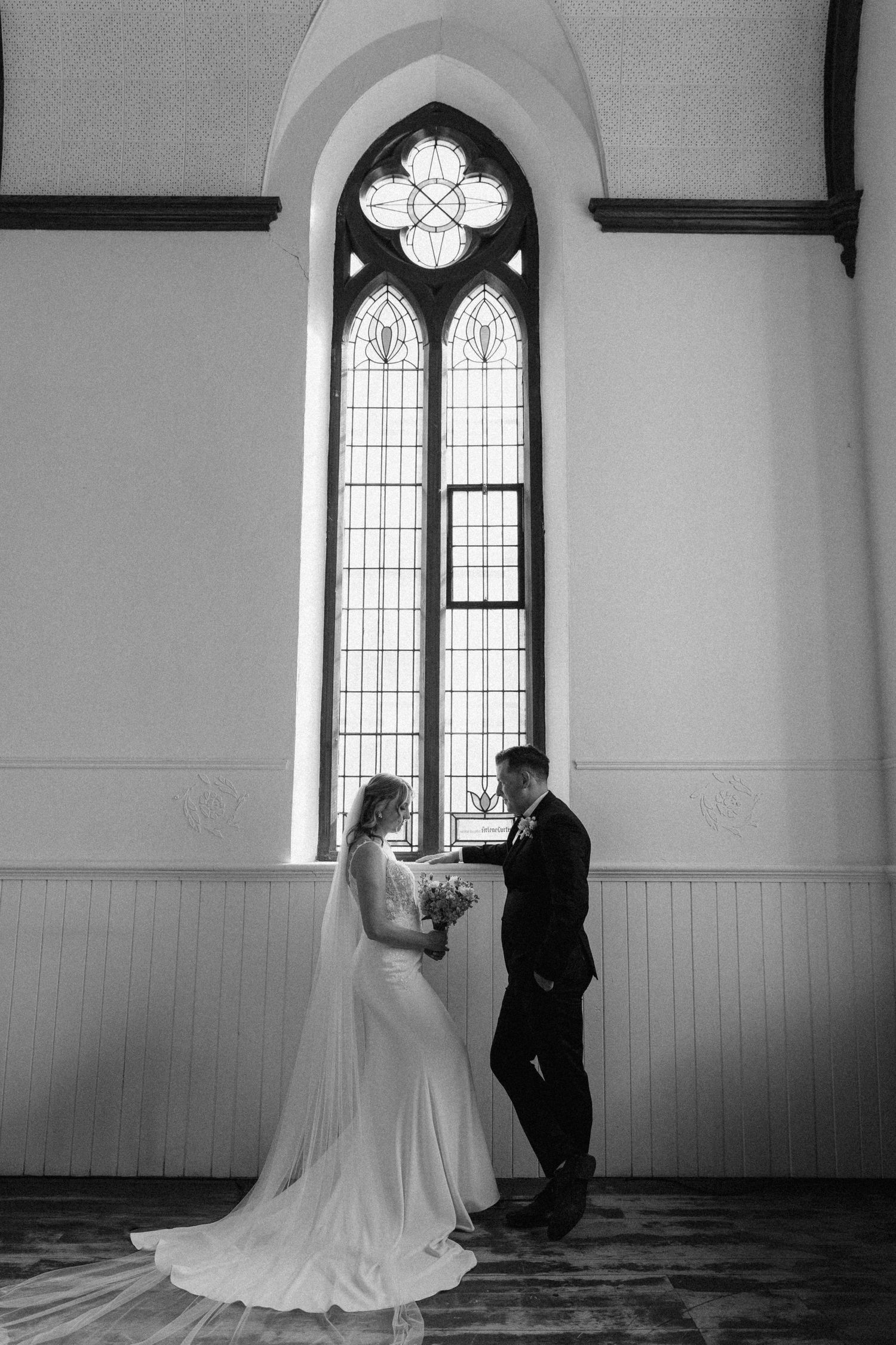 Black and white photo of bride and groom exchanging vows by a Gothic church window.