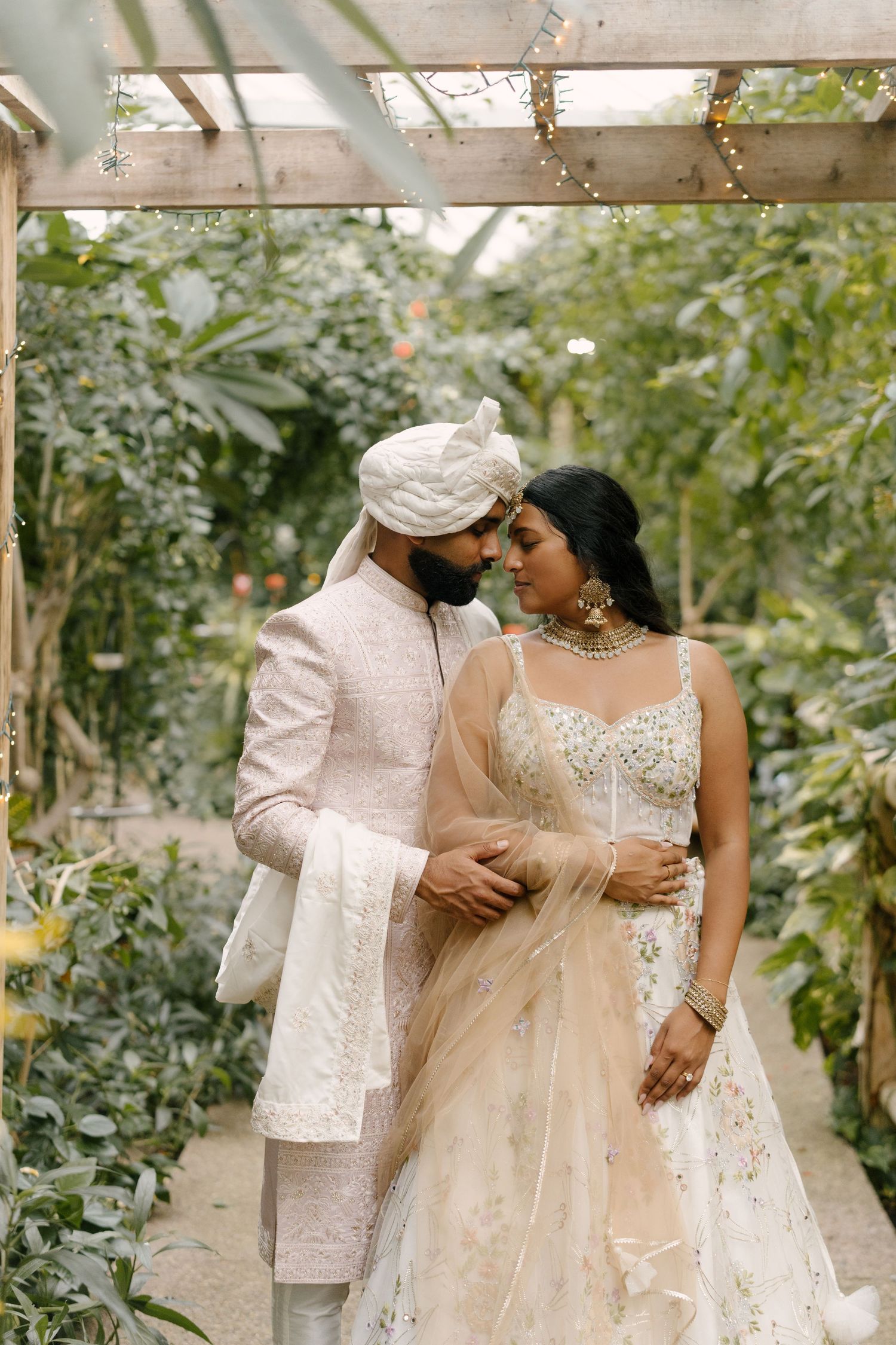 Indian couple in ivory wedding attire share a romantic moment in lush garden pergola.