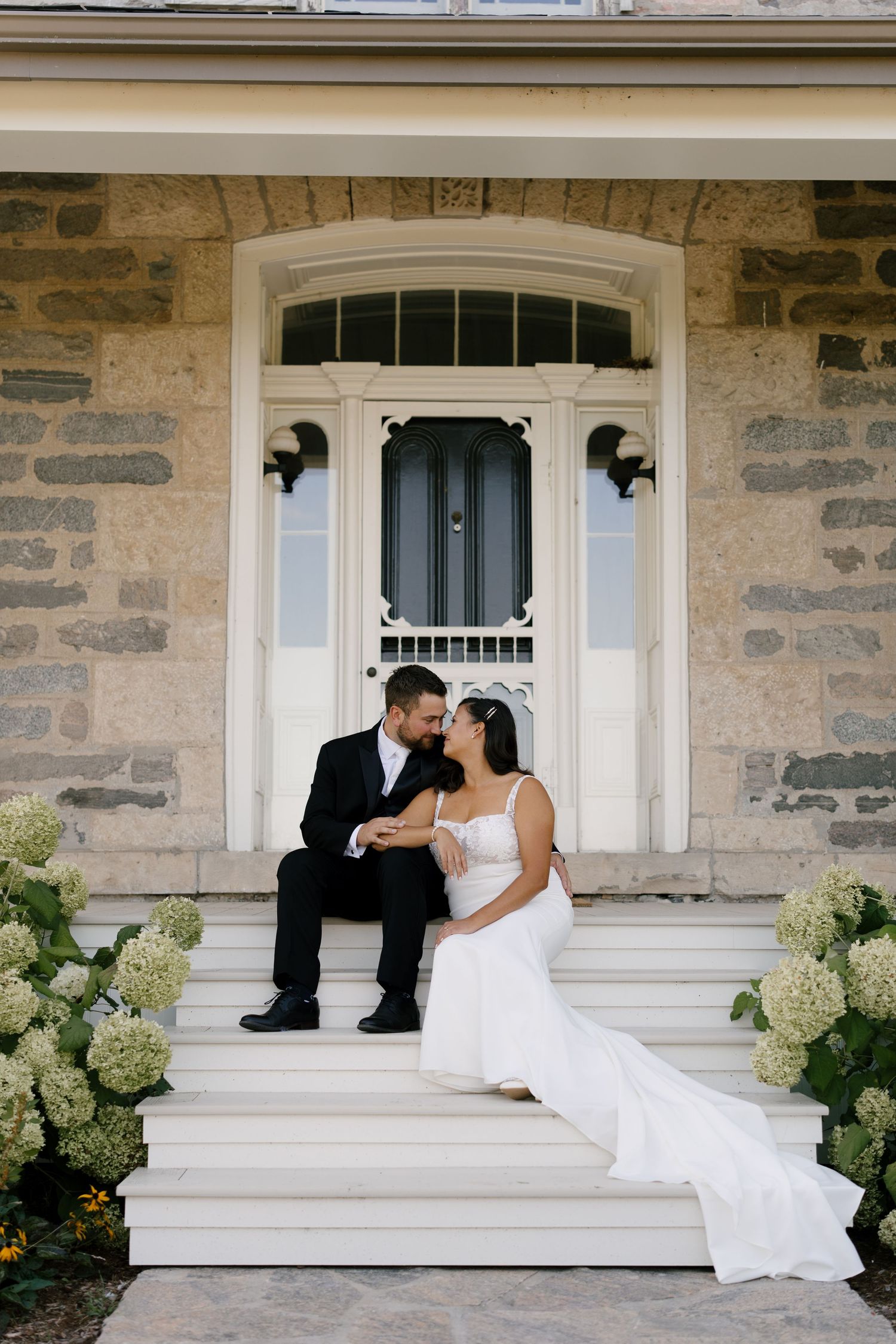 Bride and groom sit on steps of Wakefield Estates surrounded by white hydrangeas at sunset.