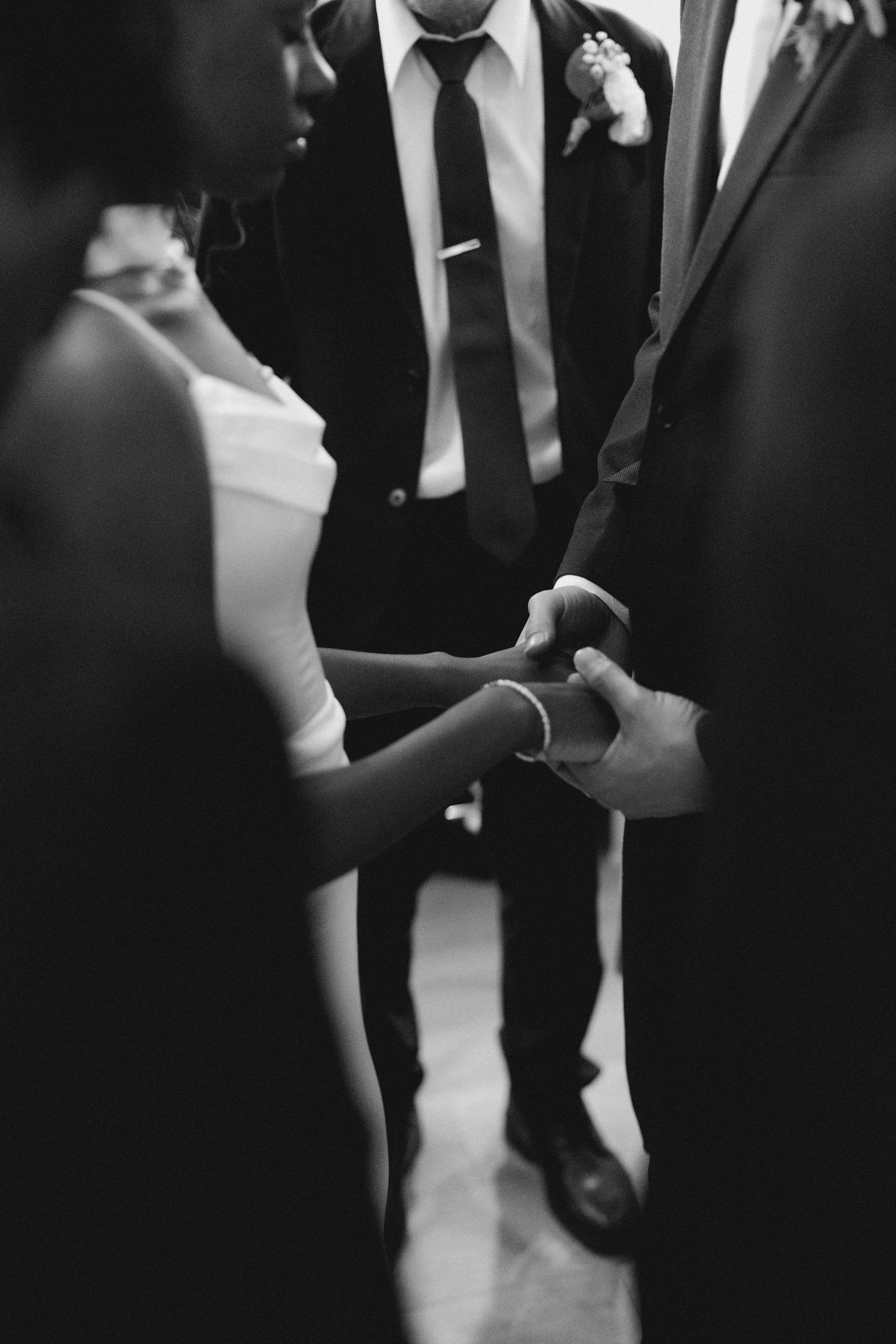Black and white photo of a couple holding hands during a wedding ceremony, with groomsmen visible in the background.