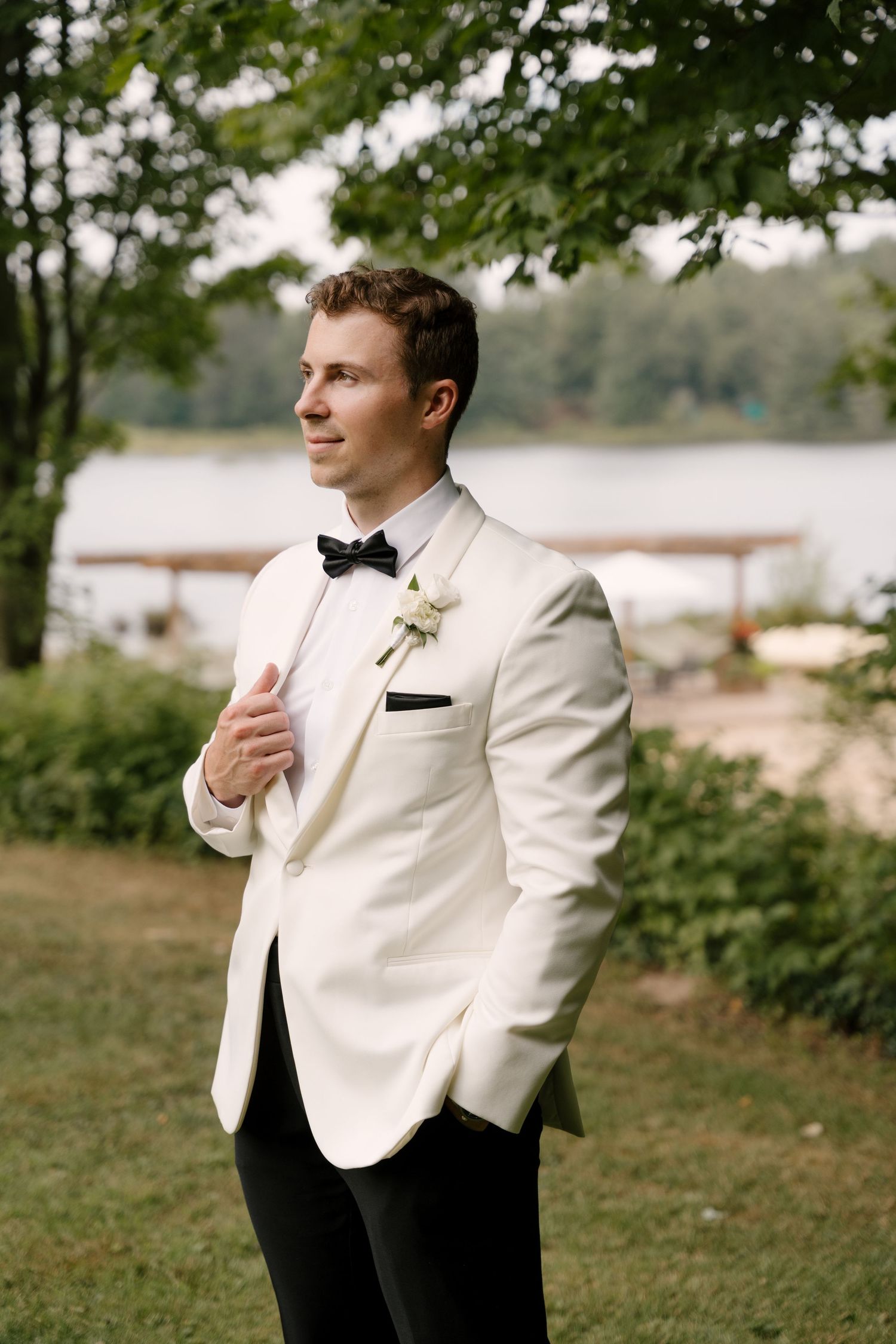 Groom in white tuxedo jacket with black bow tie and boutonniere standing outdoors near a lake.