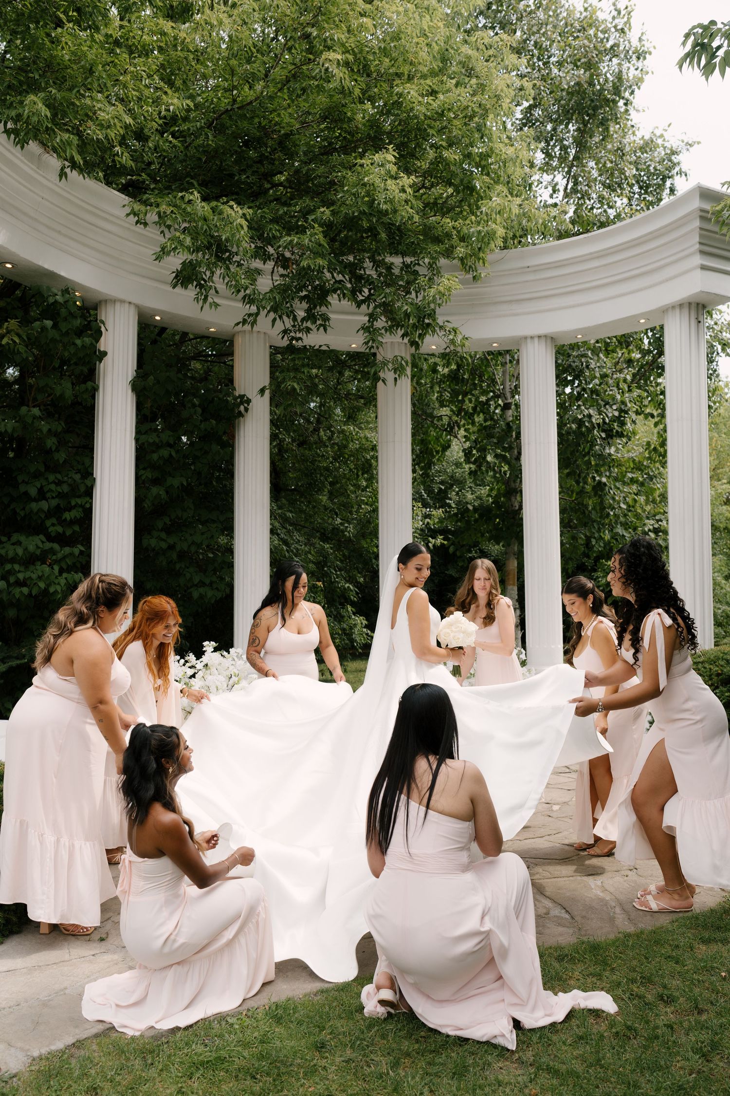 Bridesmaids in blush dresses helping arrange the bride's wedding gown train near a white columned outdoor pavilion.