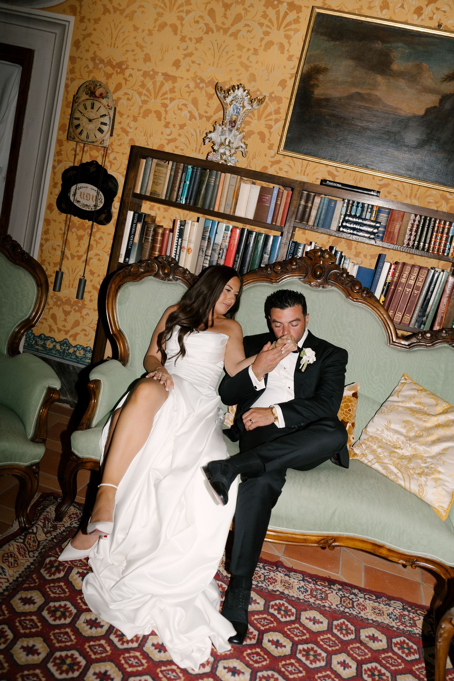 Bride and groom sitting on ornate vintage sofa in elegant library room during wedding reception.