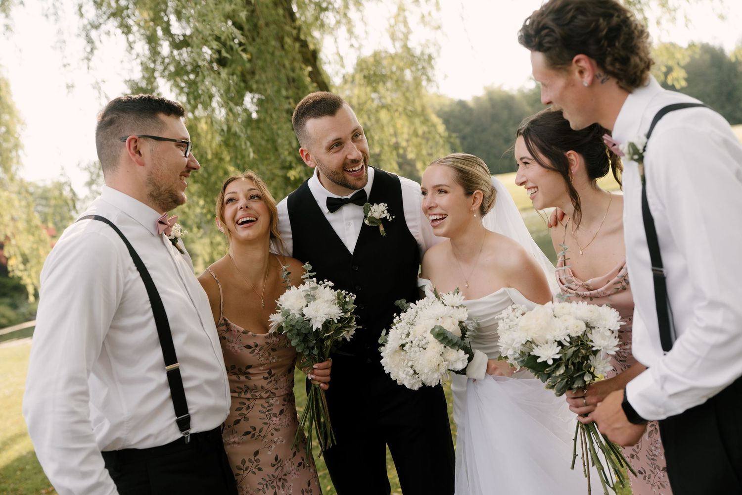 Joyful wedding party laughs together outdoors in elegant attire with white floral bouquets in Guelph.