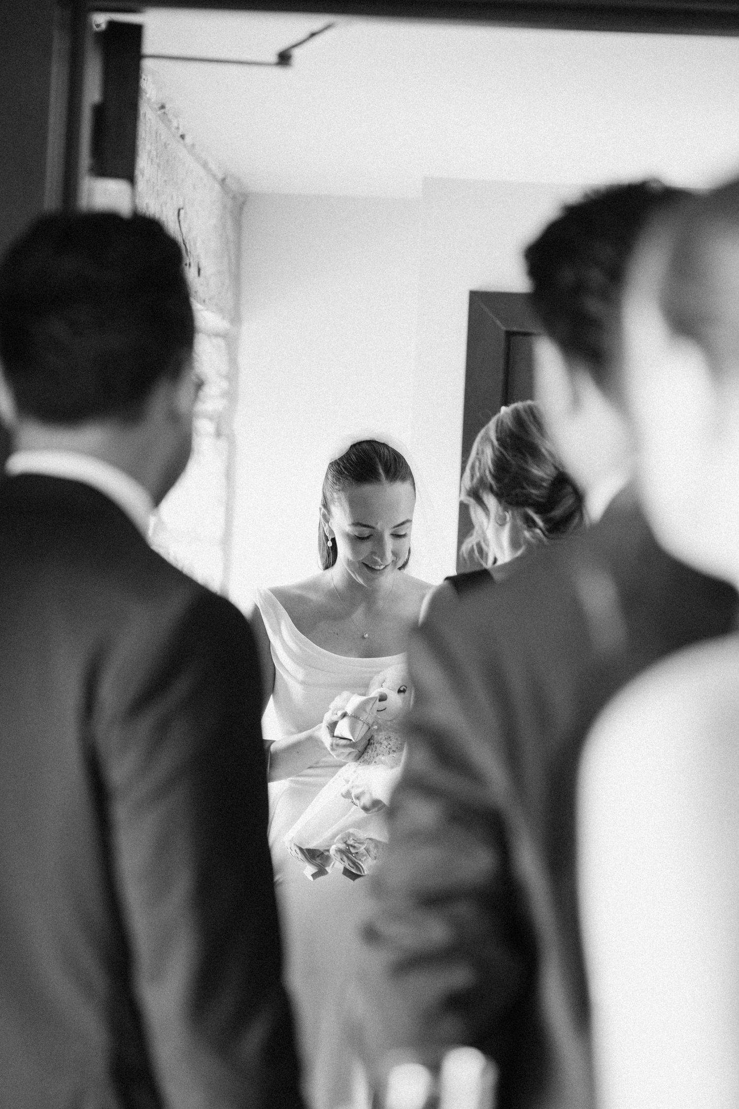 Smiling bride in white gown holds bouquet surrounded by wedding guests in black and white photo.
