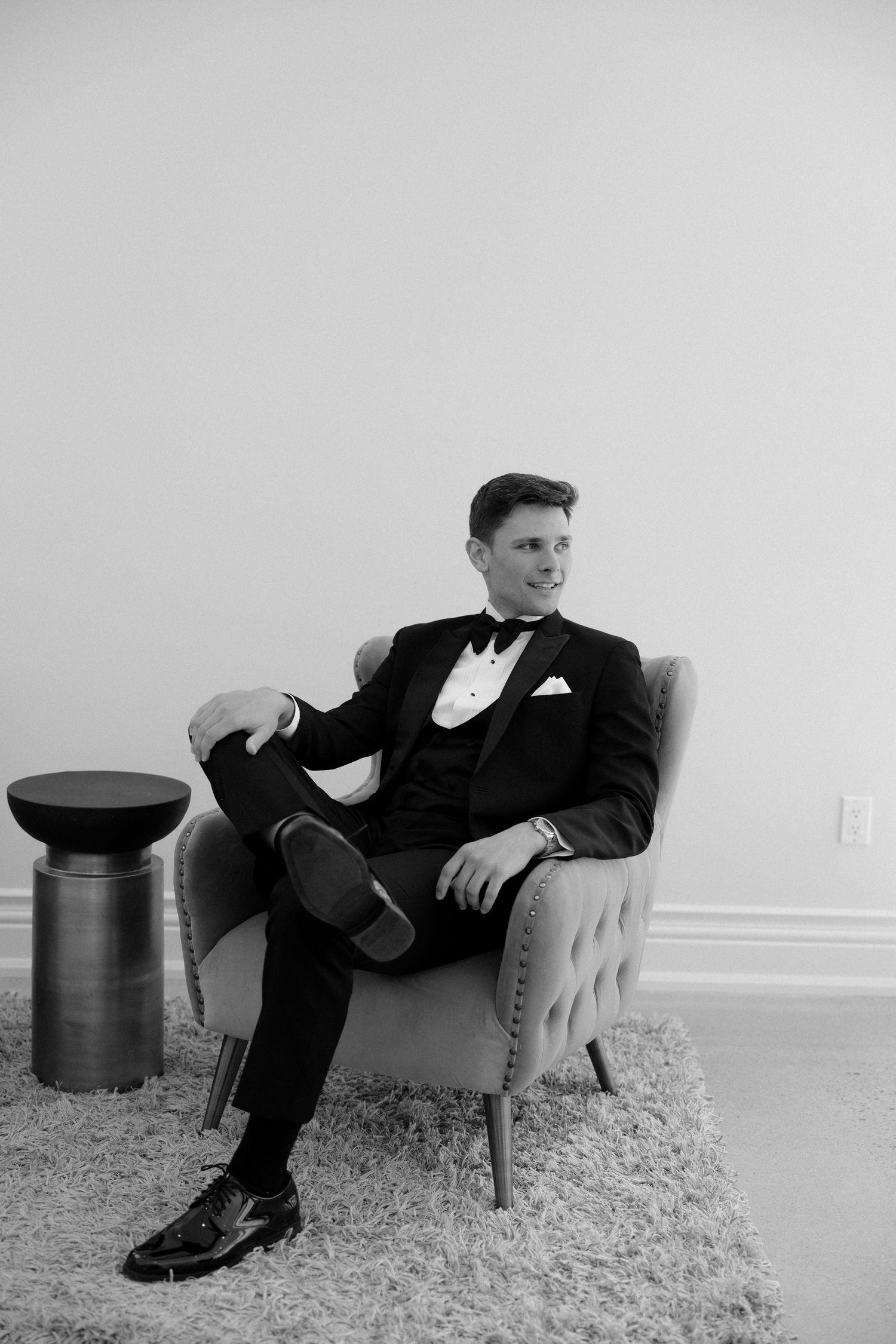 Dapper groom in a black tuxedo sitting relaxed in an armchair in a black and white photo at Roseville Estates wedding venue.