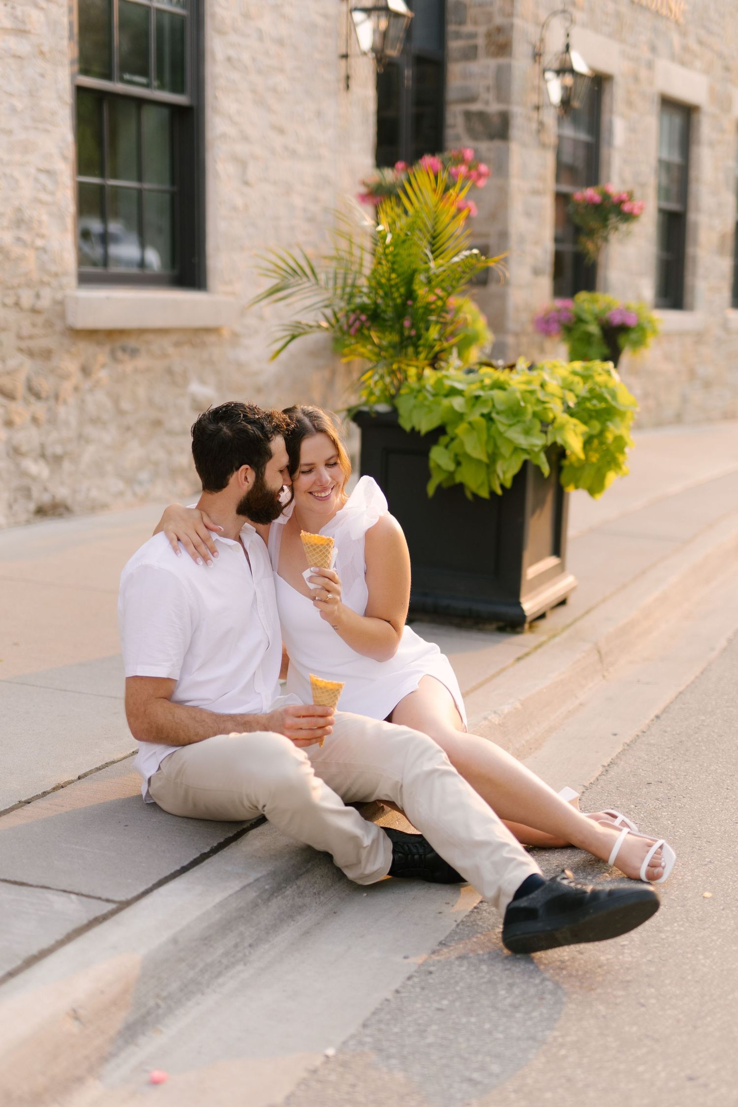 Happy couple in matching white outfits sitting on steps enjoying yellow ice cream cones together outdoors.