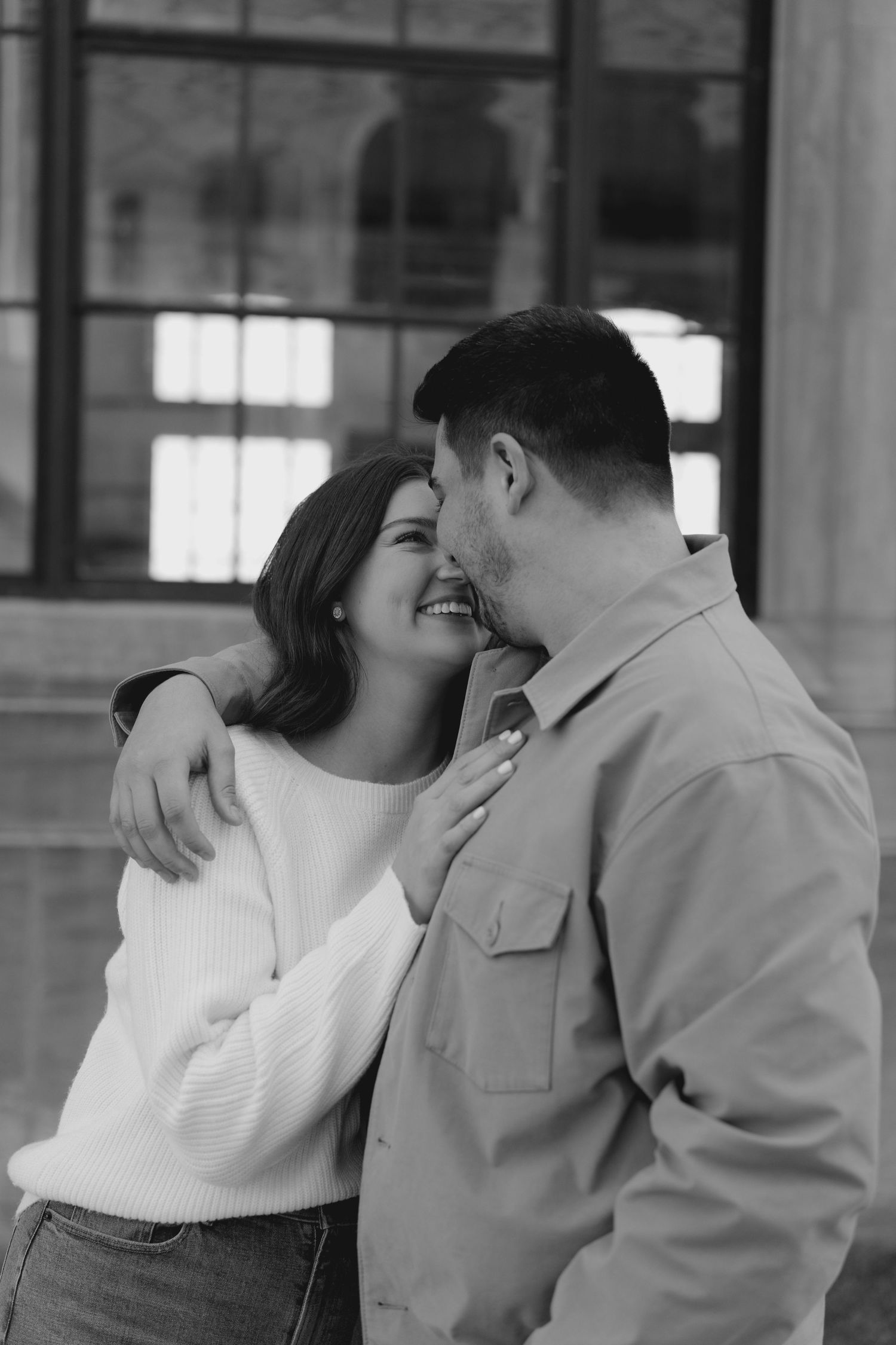 Black and white photo of laughing couple embracing outside a building with large windows behind them.