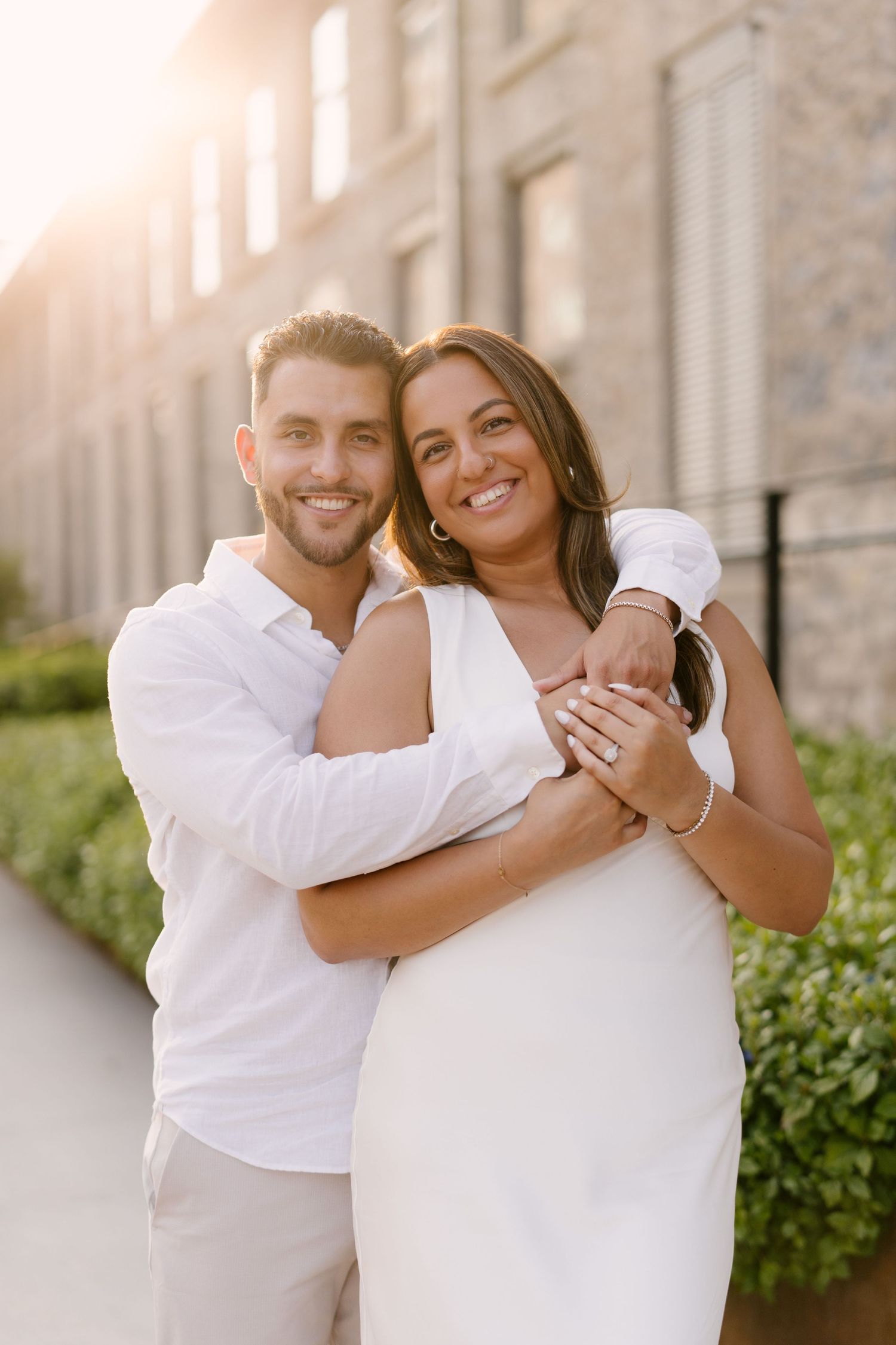 A smiling couple poses together in white outfits with golden sunlight glowing behind a stone building.