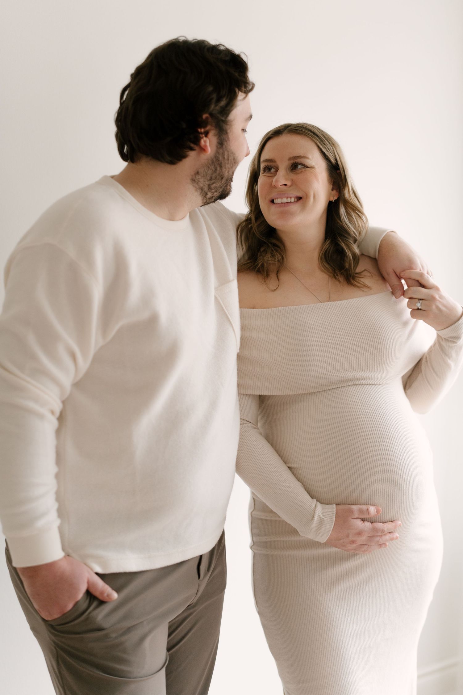 Happy pregnant couple in cream outfits smiling at each other, woman cradling her baby bump indoors.