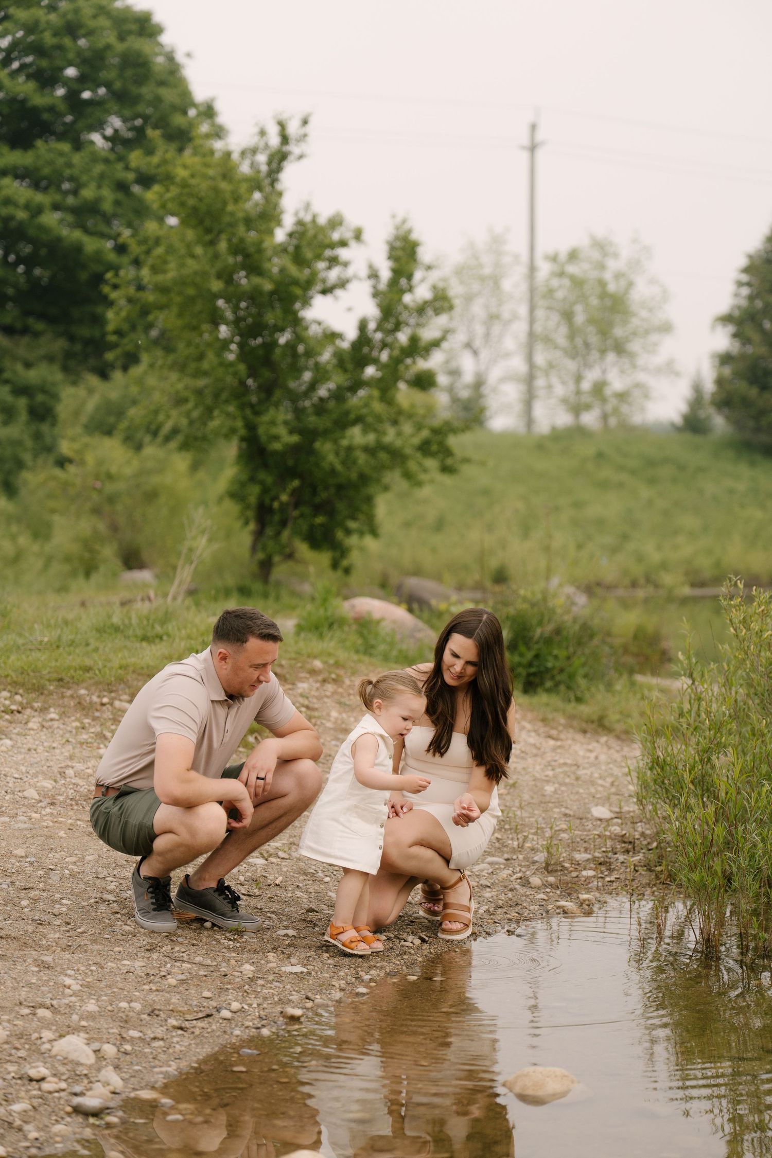 Young family crouching by a small stream, helping toddler explore nature outdoors.