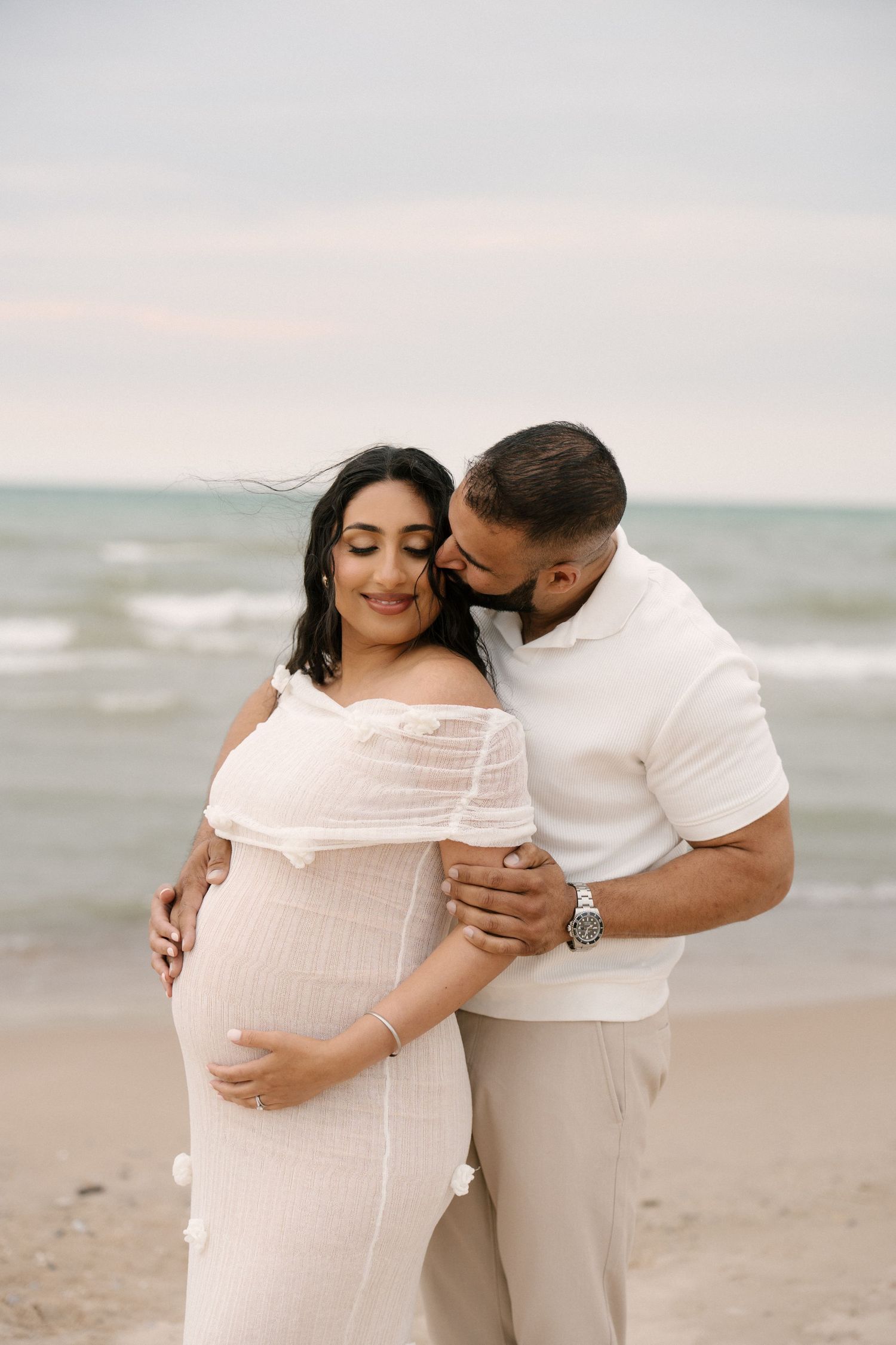 Man in white shirt kisses pregnant partner in white dress on beach, both cradling her baby bump.