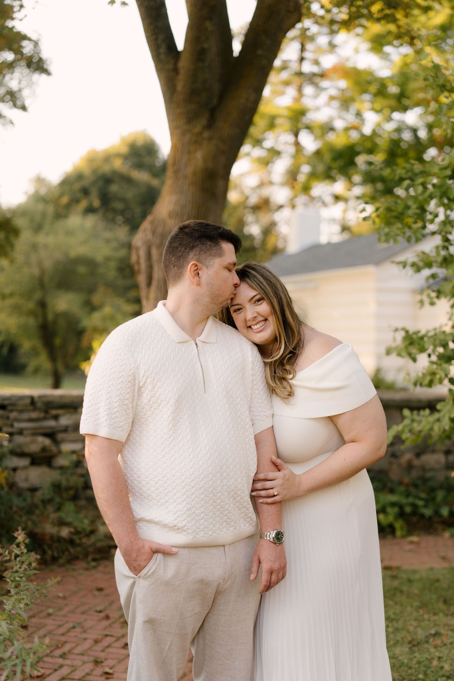 Couple in white outfits embracing outdoors near stone wall and trees during golden hour.