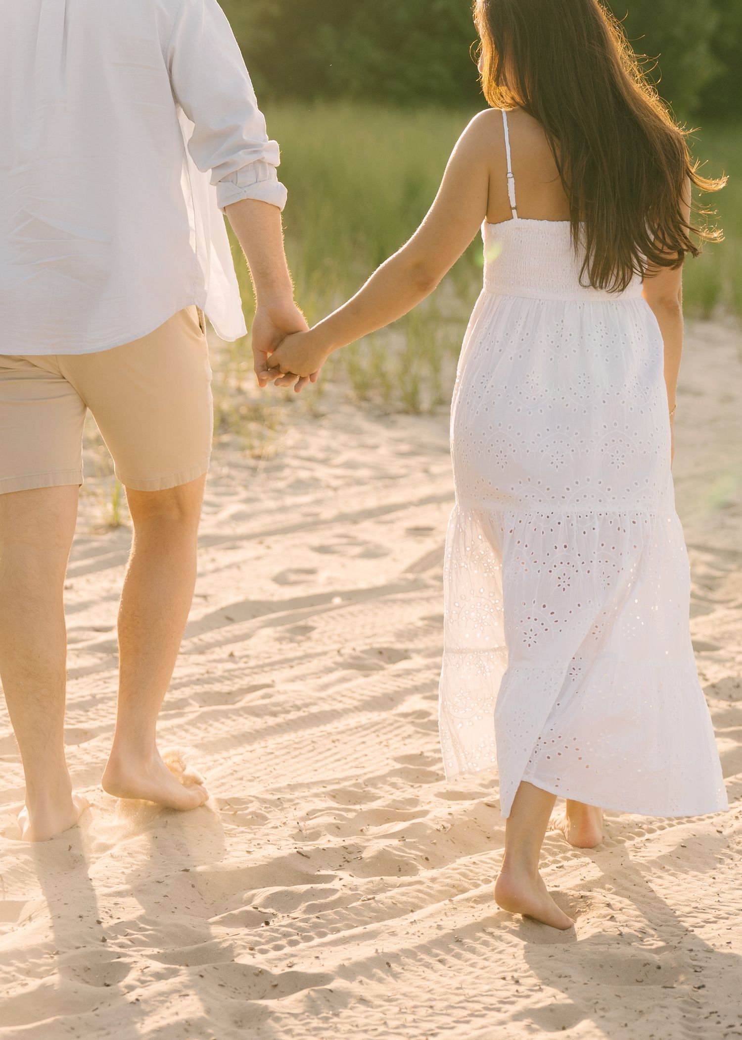 Couple holding hands walking barefoot on sandy beach, woman in white eyelet maxi dress, man in white shirt and tan shorts.