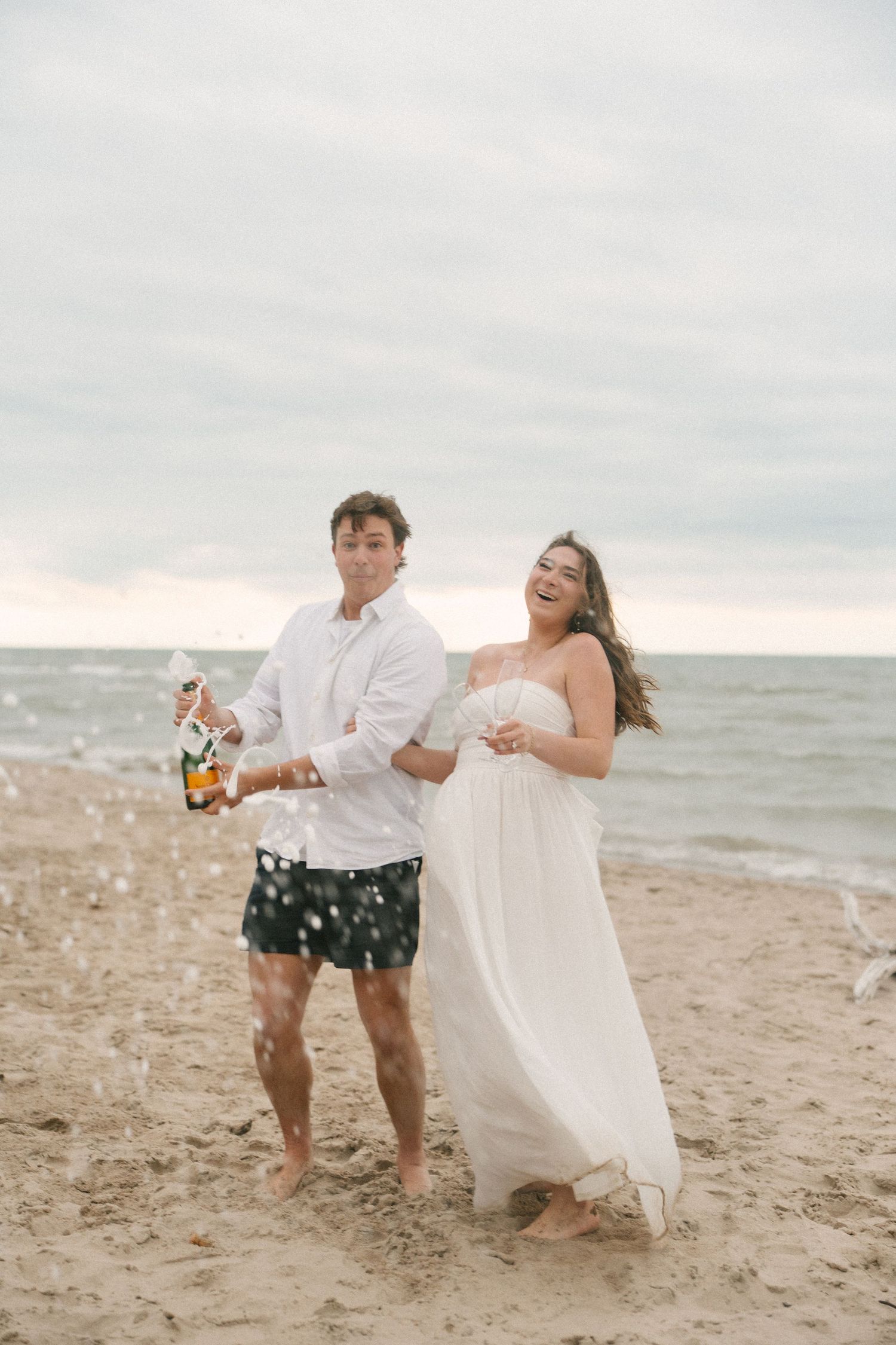 Joyful couple popping champagne bottle on sandy beach, woman in white dress laughing as bubbles spray in air.