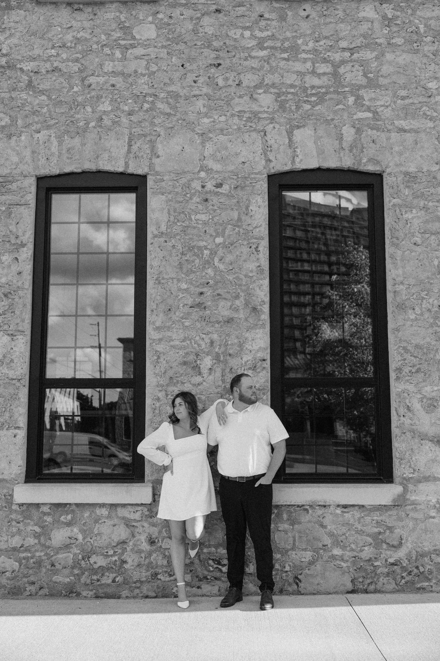 Black and white photo of couple standing apart before a stone wall with large windows.