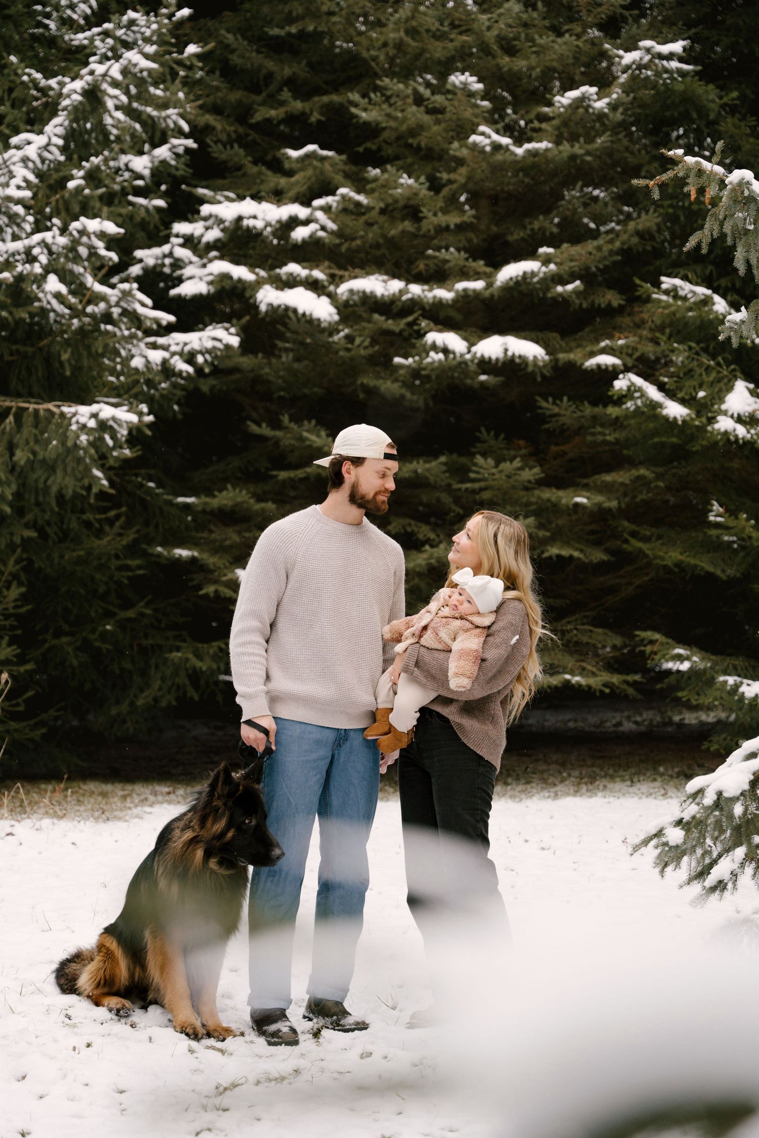 Young family with newborn baby and German Shepherd dog posing in snowy pine forest in winter.