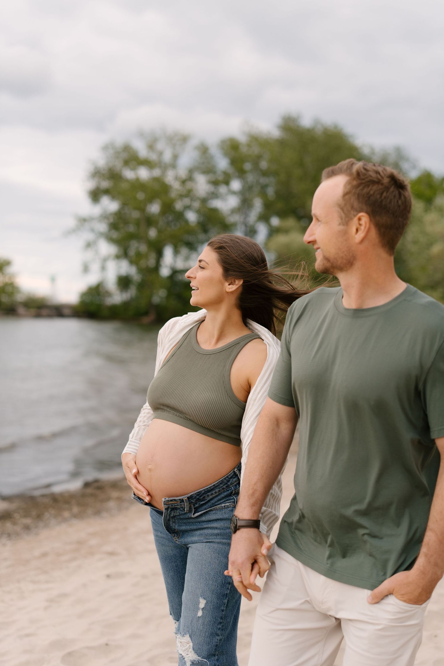 Pregnant woman in olive crop top and jeans holds her baby bump while walking hand-in-hand with partner on sandy beach.