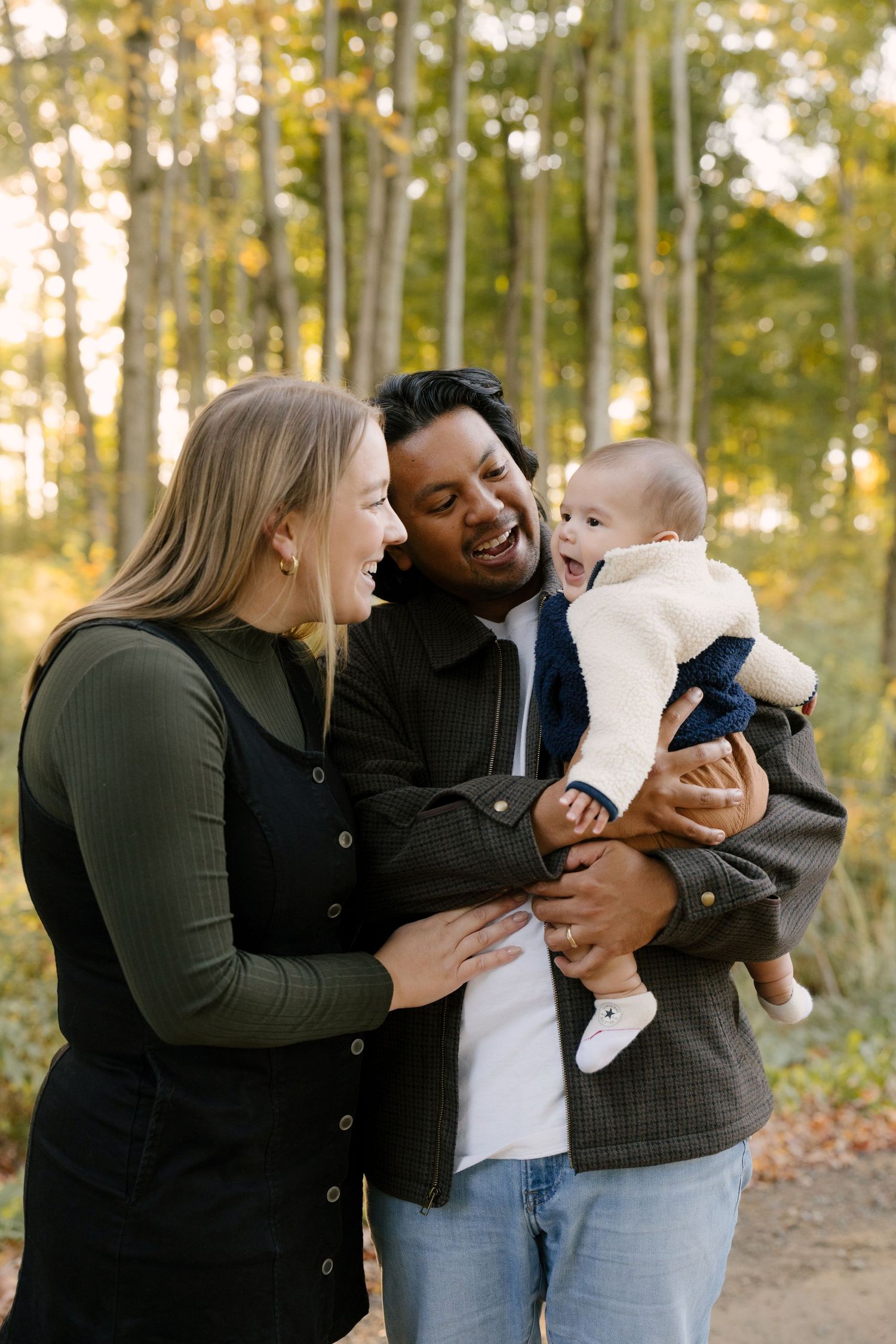 Laughing interracial couple in autumn forest, man holding giggling baby in white sweater.