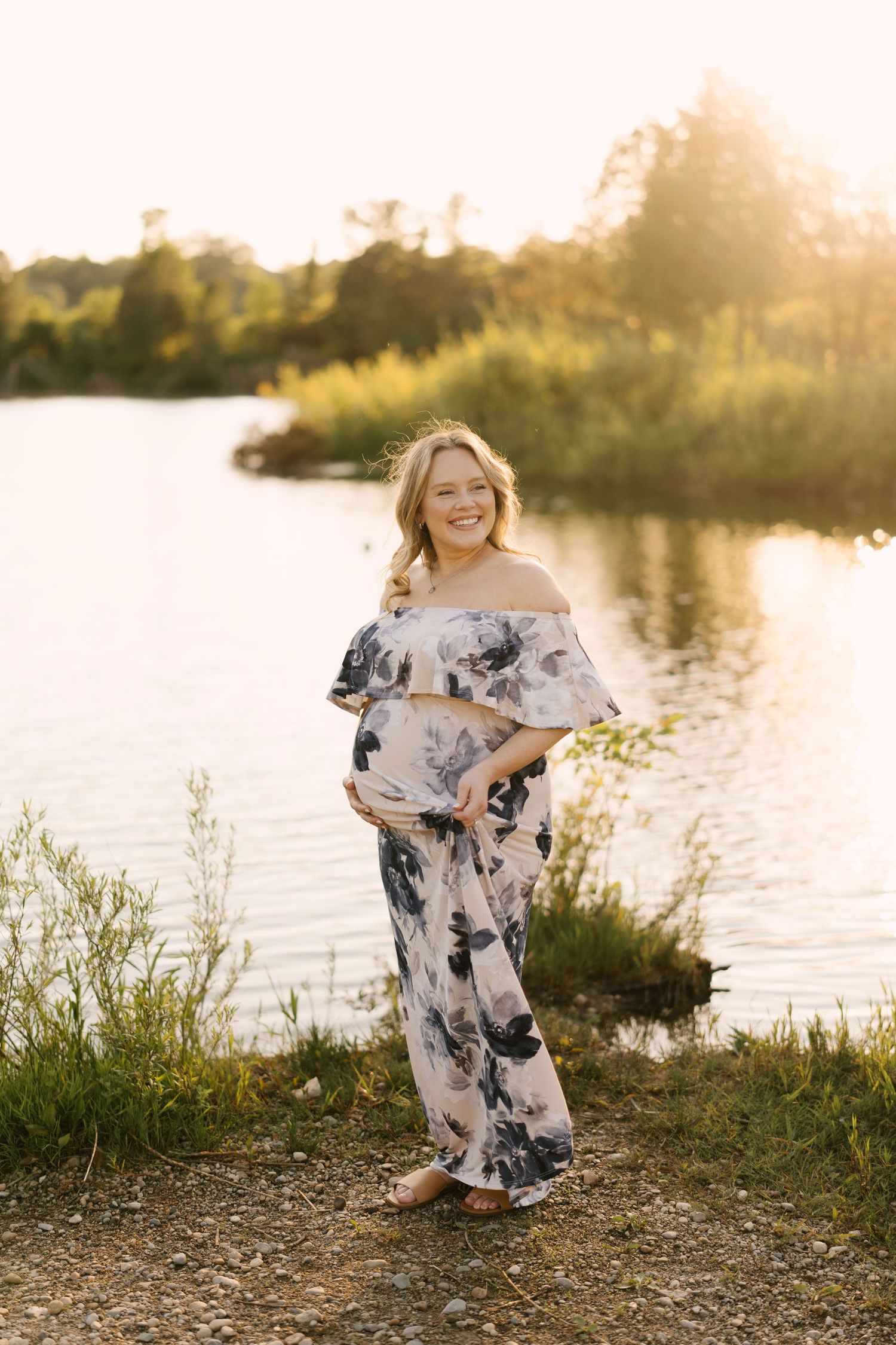 Smiling pregnant woman in off-shoulder floral maxi dress stands by a reflective lake at golden sunset.