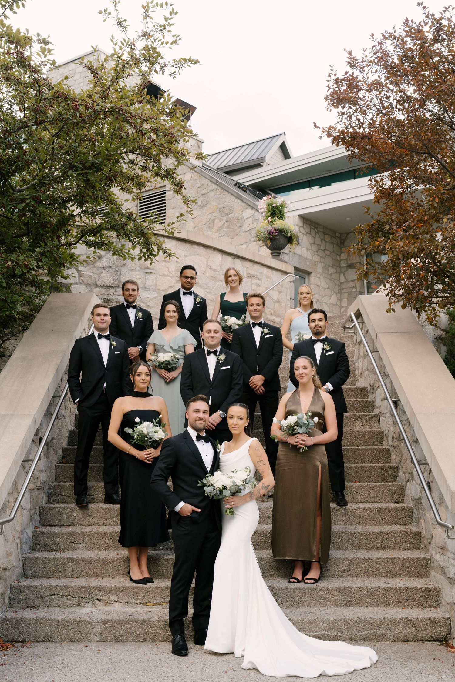Wedding party posing on stone staircase outside elegant venue in classic black and white attire at Cambridge Mill.