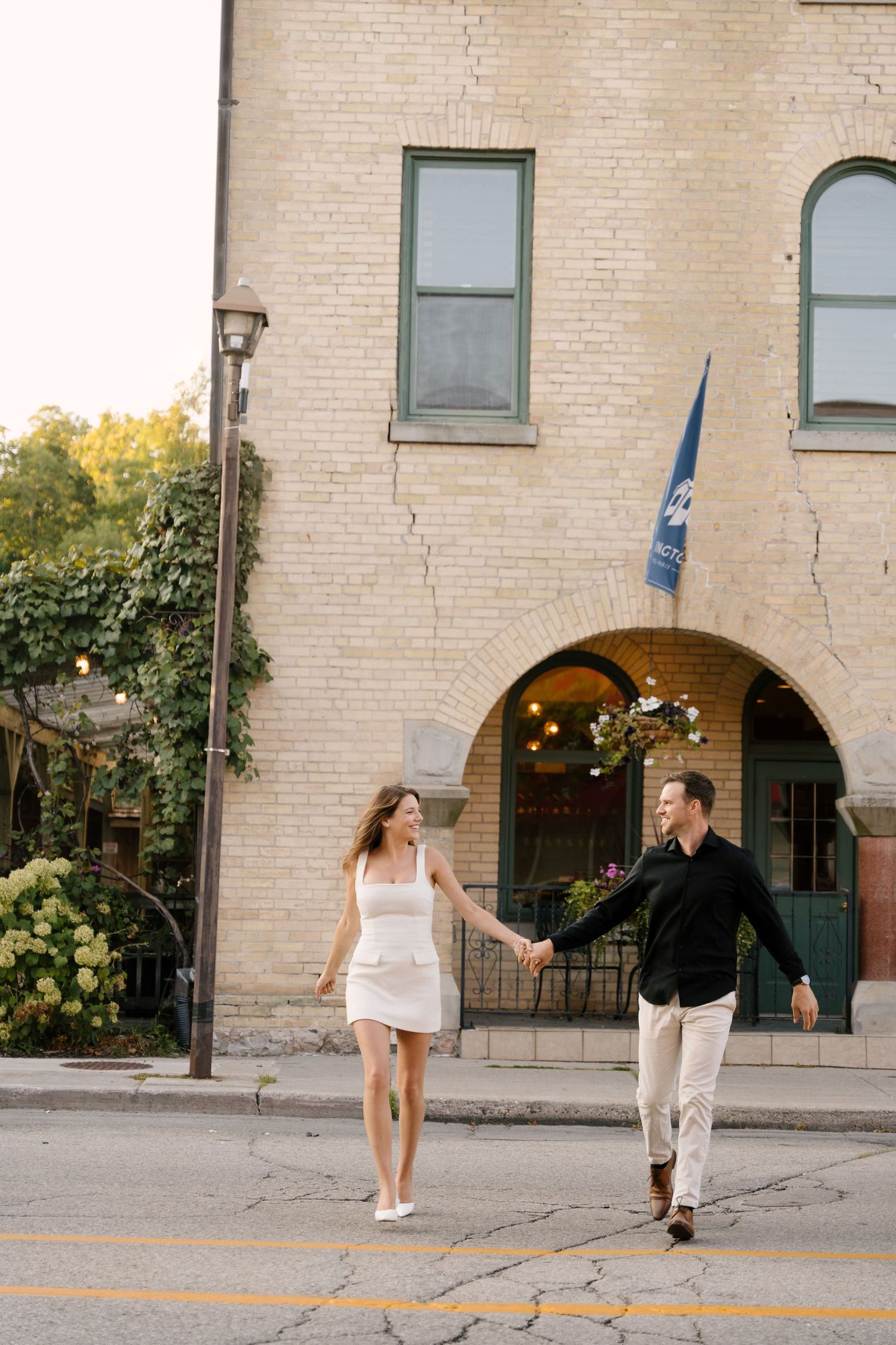 Stylish couple holding hands crossing a street in front of a charming historic brick building in a quaint town.