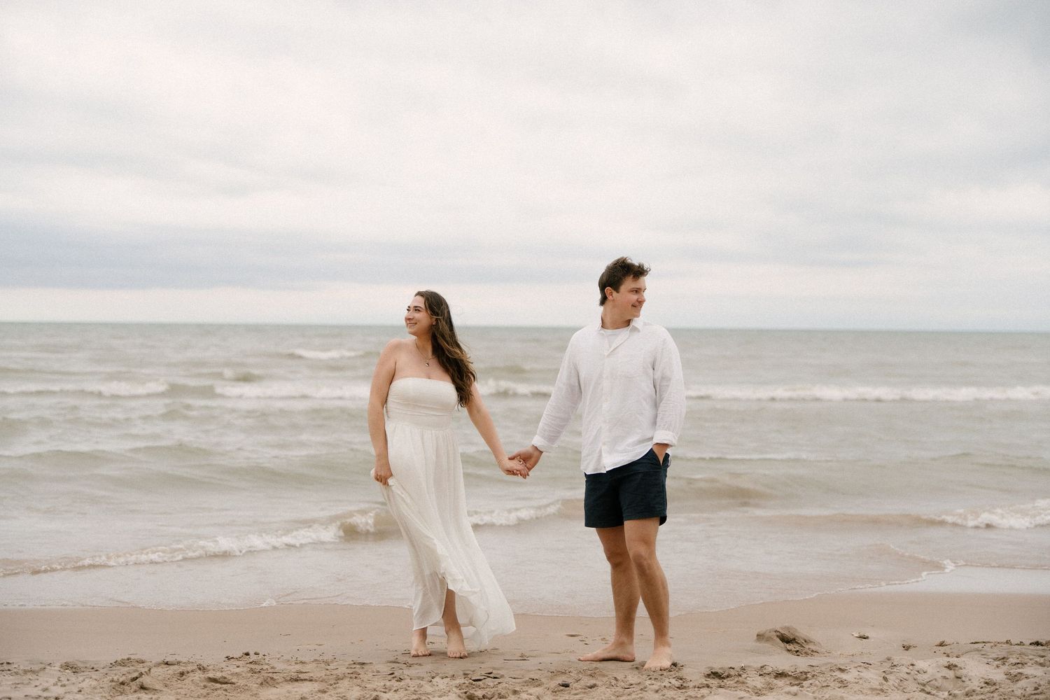 Young couple holding hands and walking barefoot on a sandy beach with overcast skies and ocean waves behind them.
