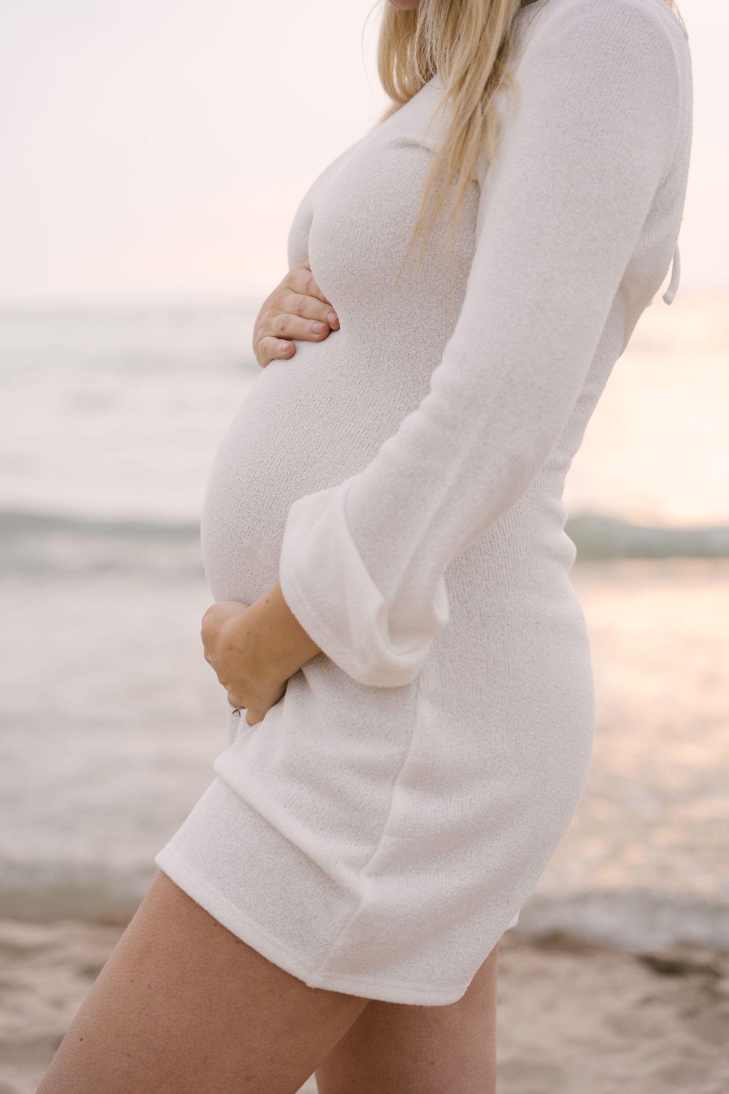 Pregnant woman in white knit dress cradling baby bump on beach at sunset.