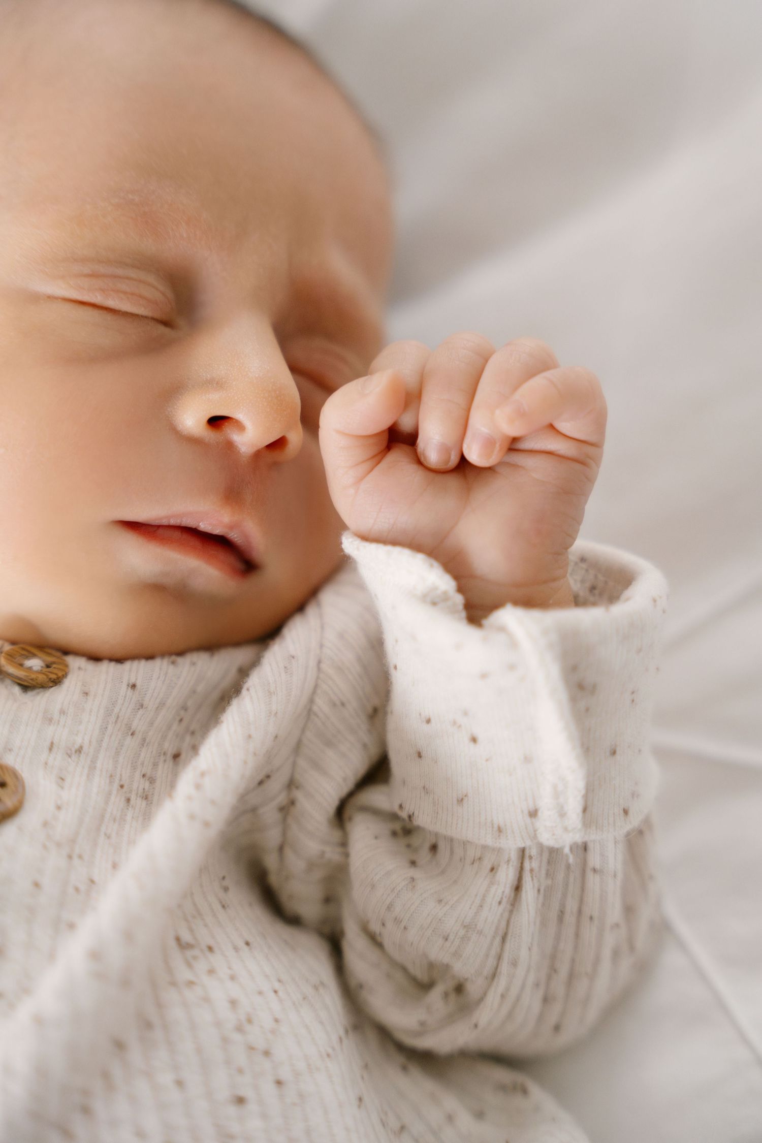 Close-up of sleeping newborn baby in cream knit cardigan with tiny fist near face.