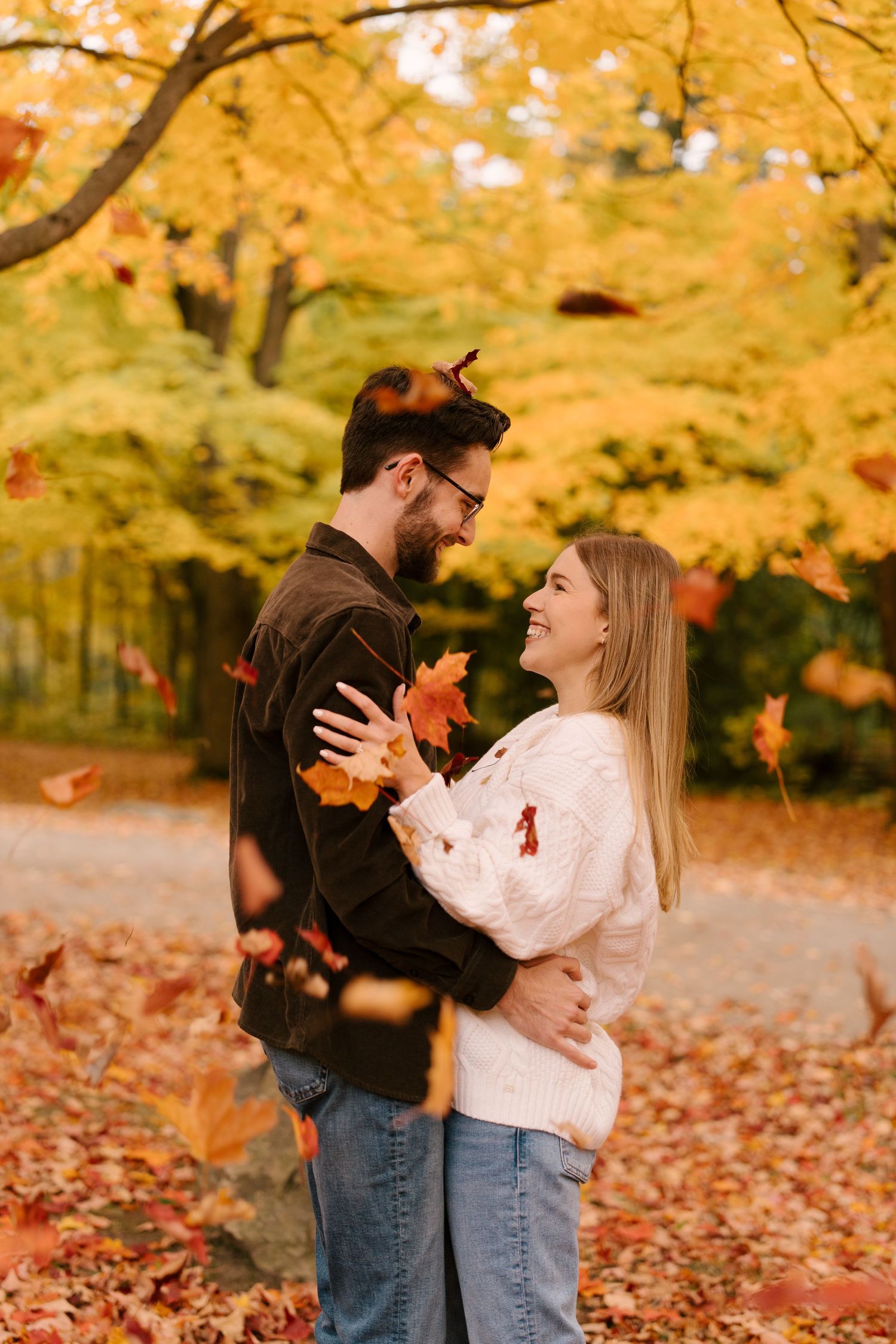 A laughing couple embraces as colorful autumn leaves fall around them in a golden forest.