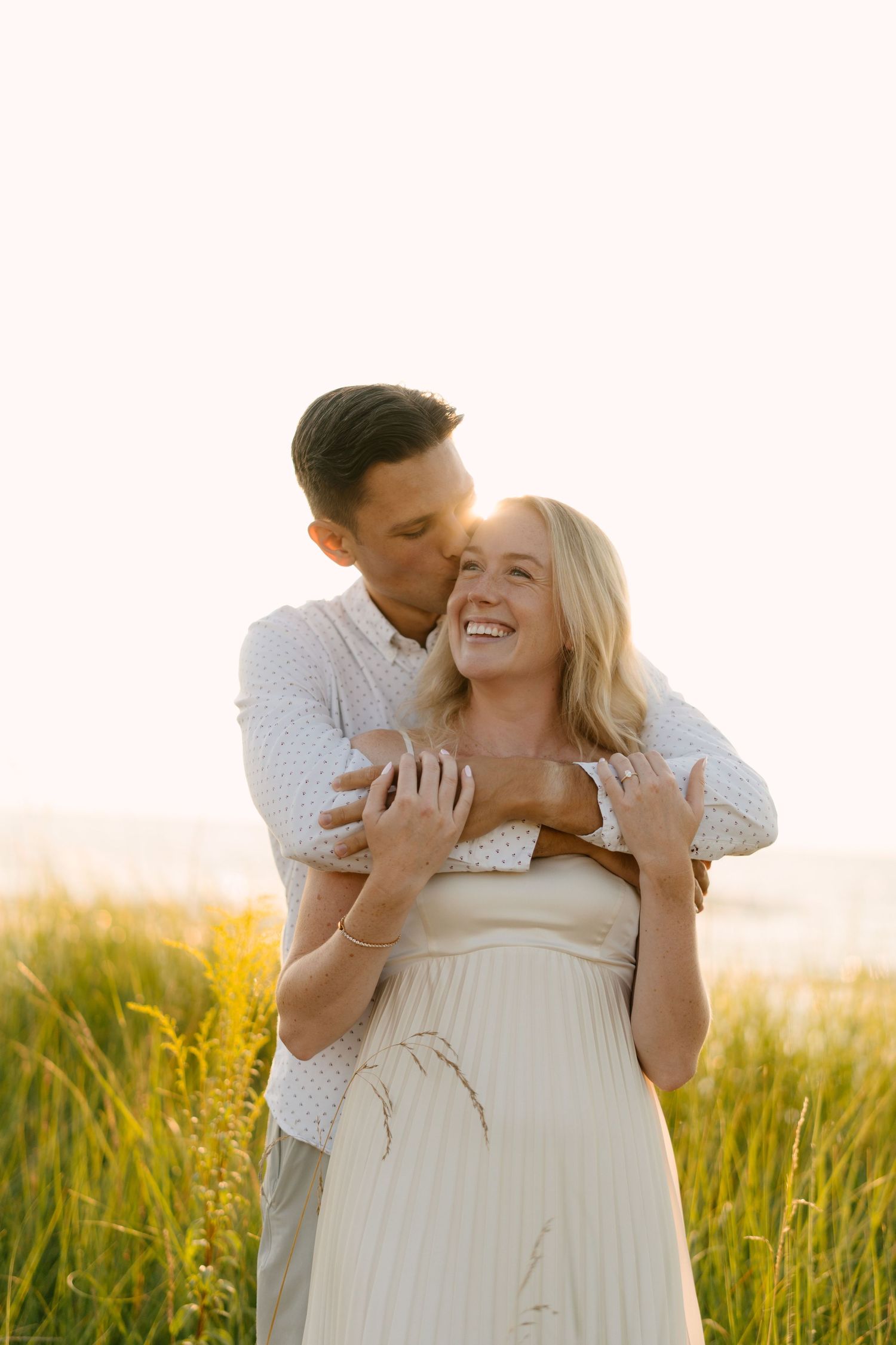 Man kissing woman on cheek from behind at golden hour, both laughing near lush beach grass.