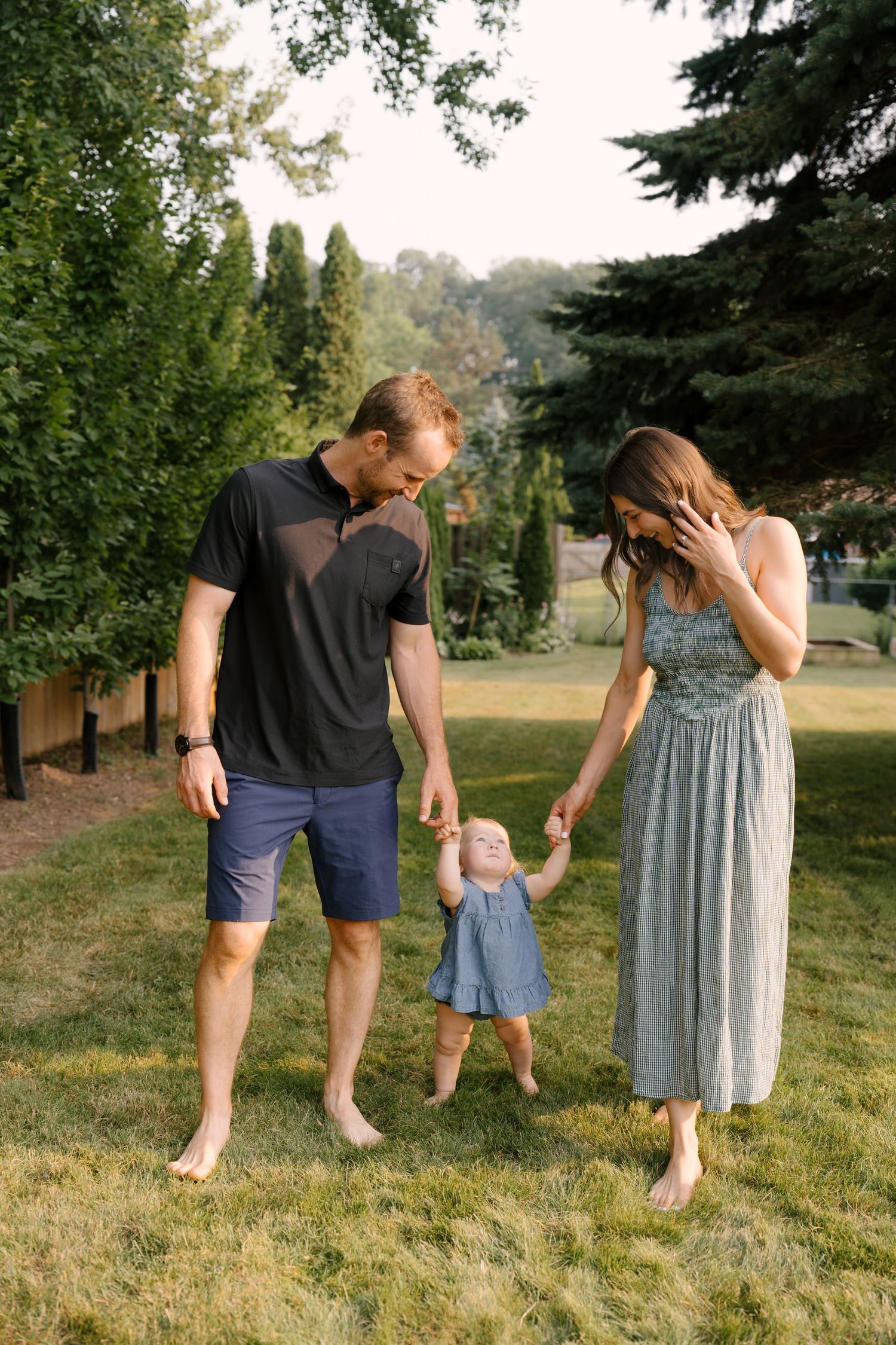 Barefoot parents hold their toddler daughter's hands as she takes steps on a lush green lawn.