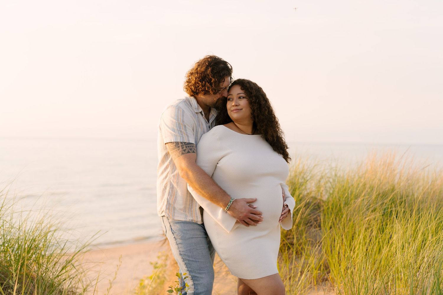 Pregnant woman in white dress embraced by partner on a beach at sunset.