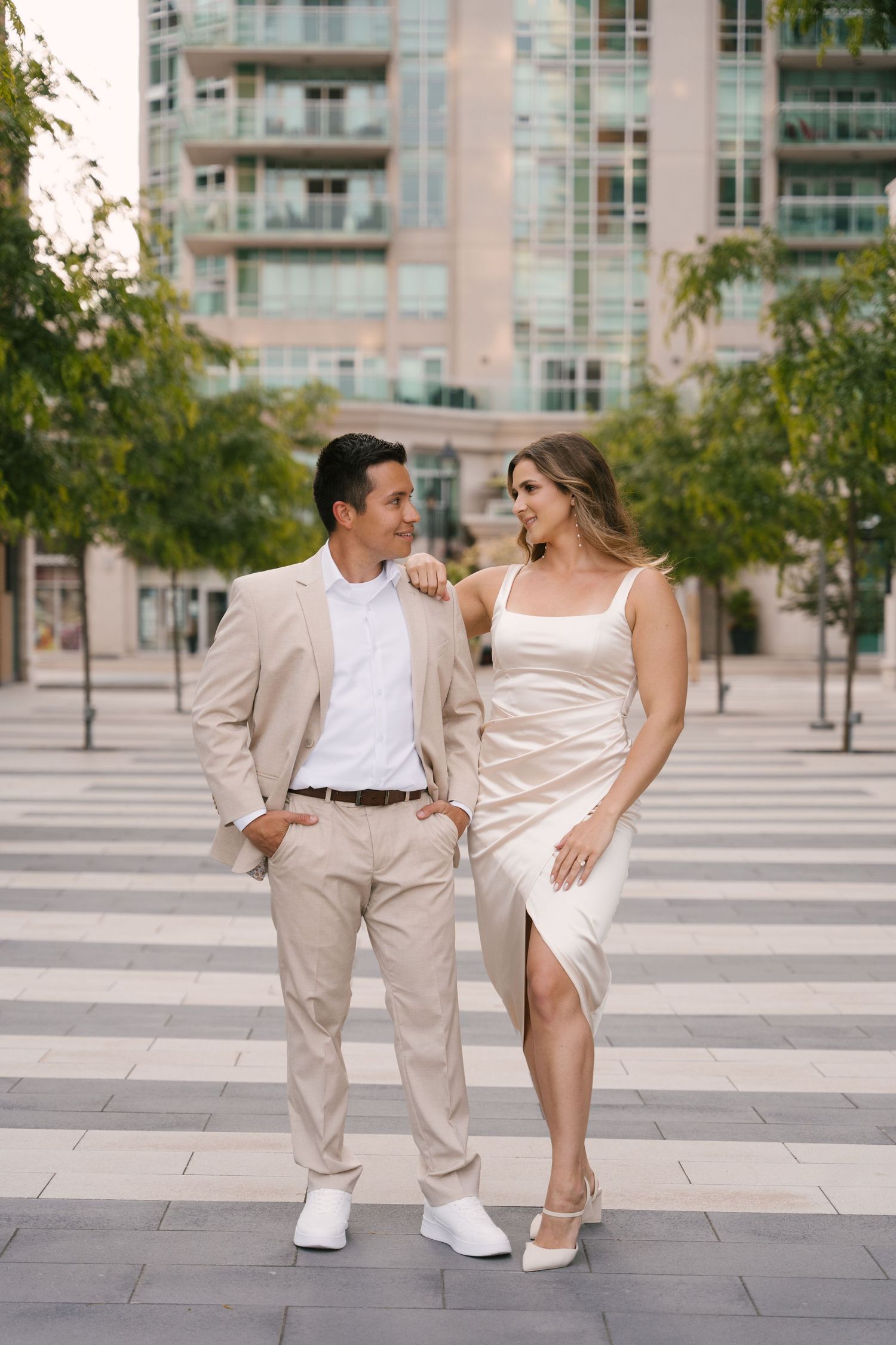 Stylish couple in coordinating beige outfits posing together on an urban plaza with modern buildings.