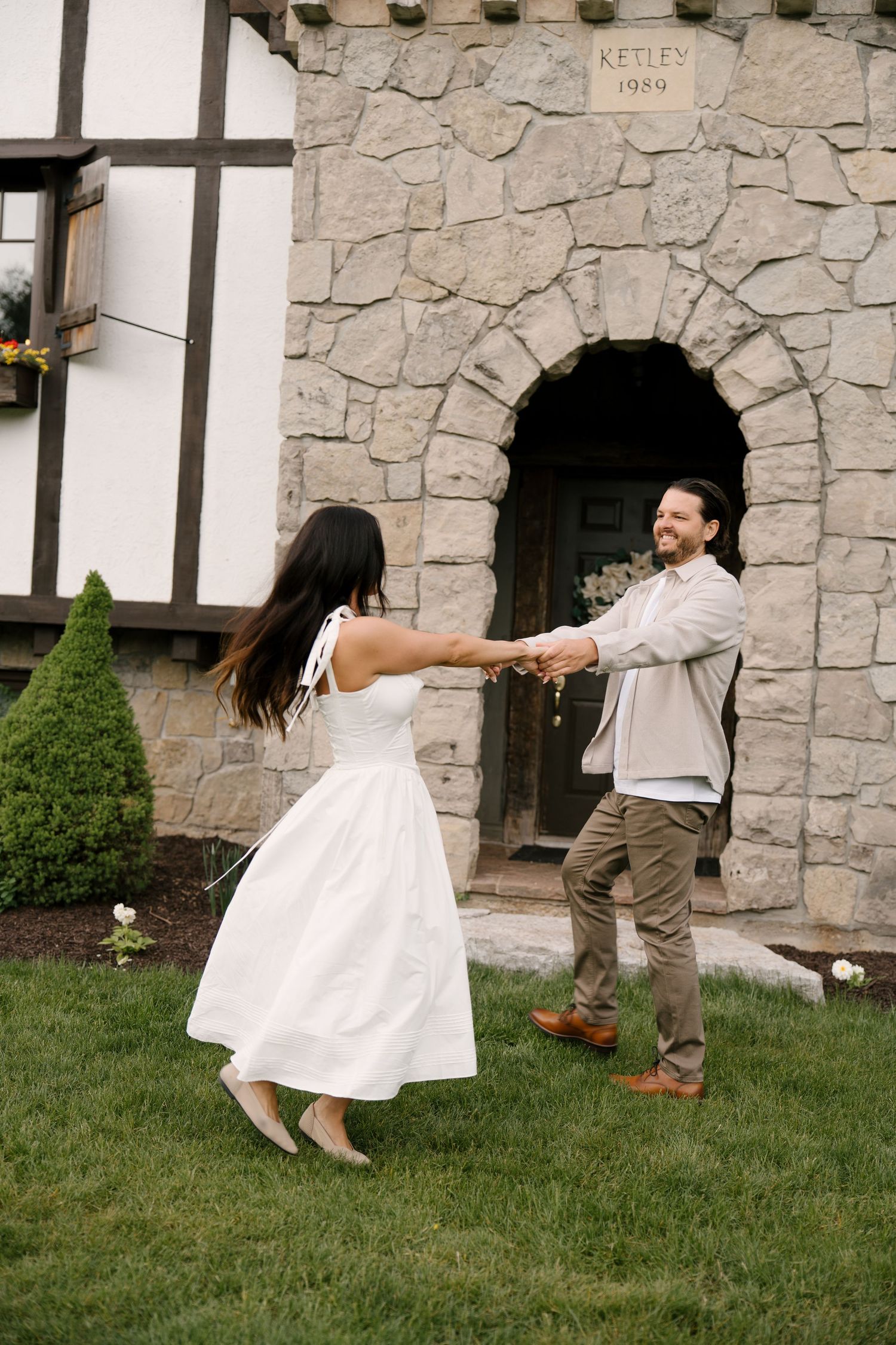 Couple dancing and holding hands on green lawn in front of stone arch building entrance.
