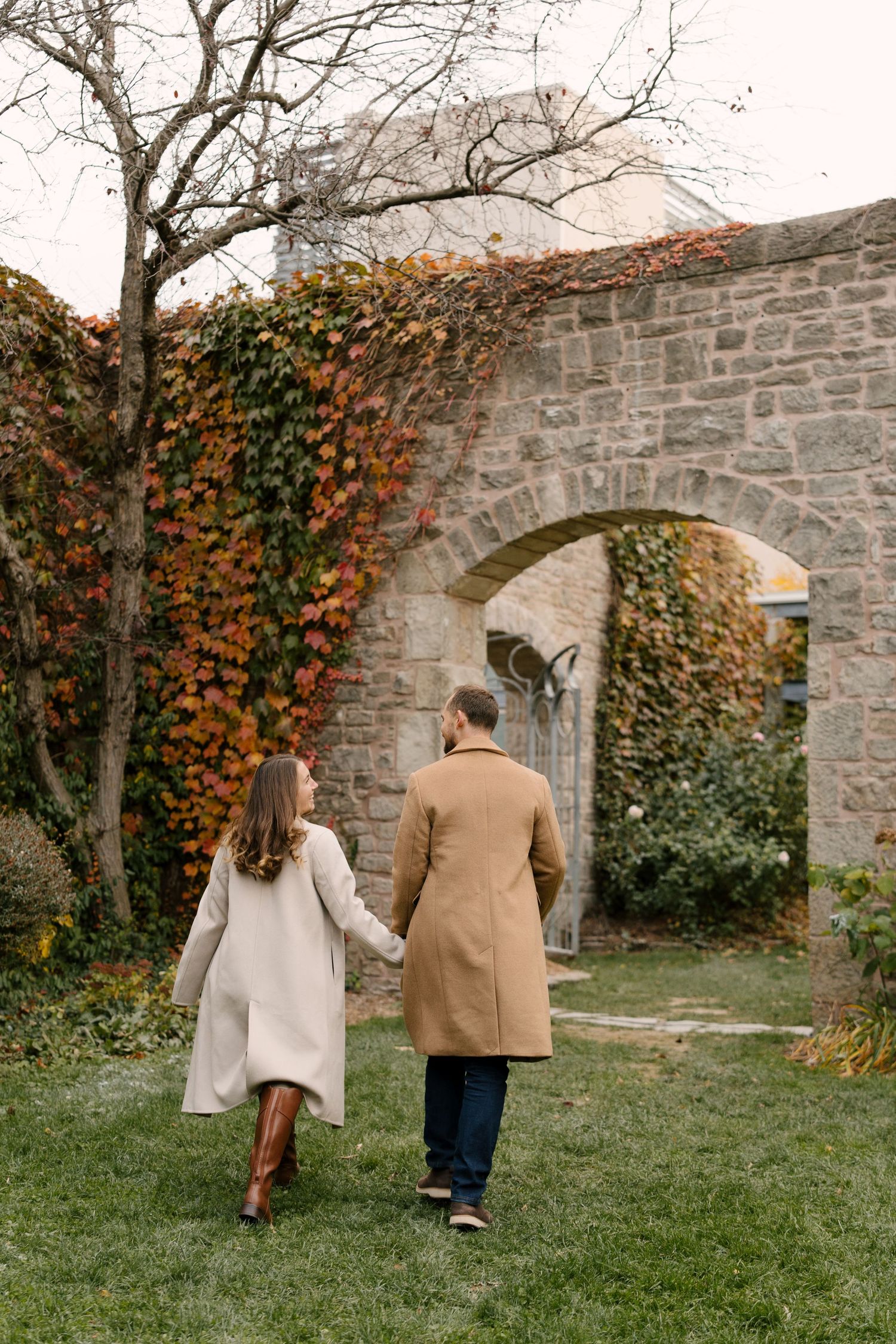 Couple holding hands walking toward a stone archway covered in vibrant autumn ivy in a garden.