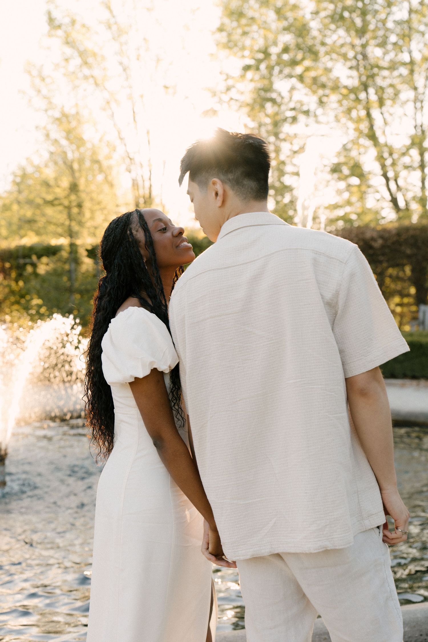 Couple holding hands facing each other near a garden fountain in warm golden sunset light.