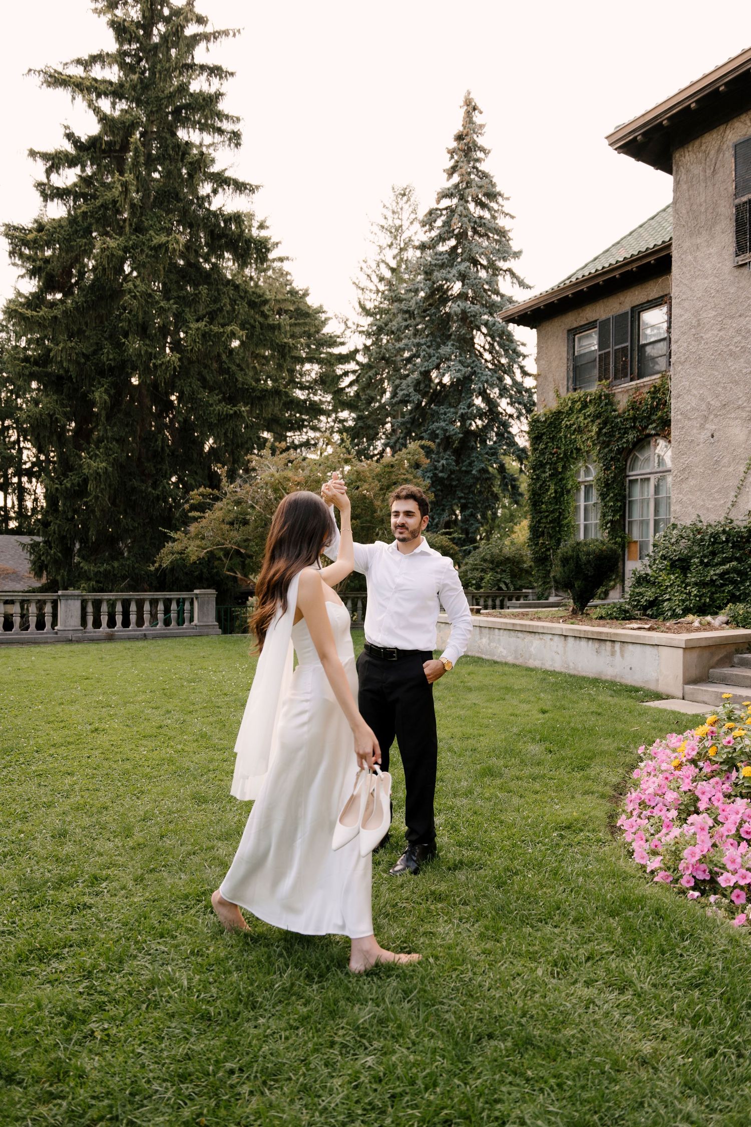 Joyful couple dancing barefoot on green lawn beside ivy-covered estate with tall evergreen trees in background.