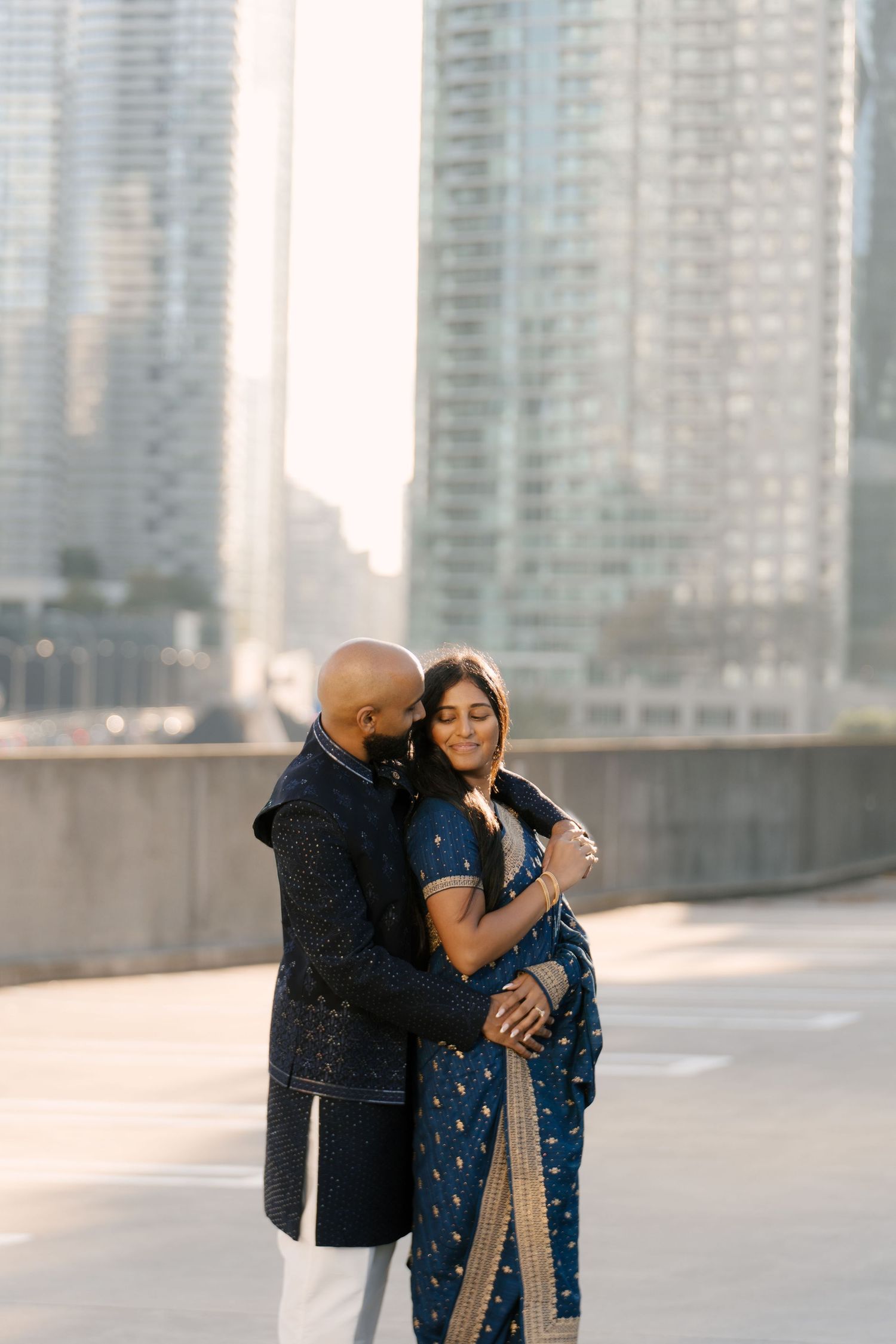 Indian couple in blue traditional wear share a romantic embrace with city buildings behind.