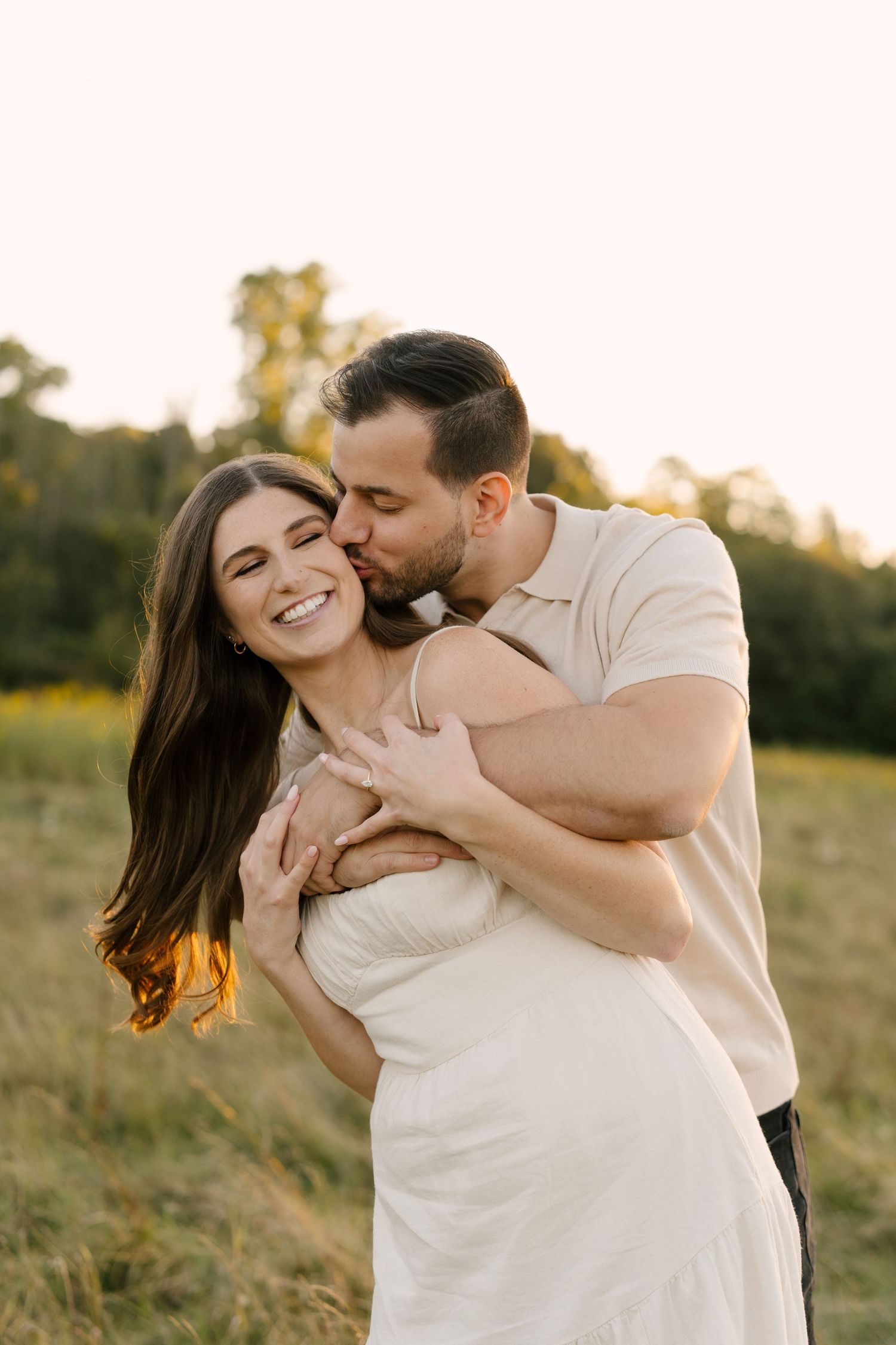 Man kissing smiling woman on the cheek while embracing her in a sunlit meadow.