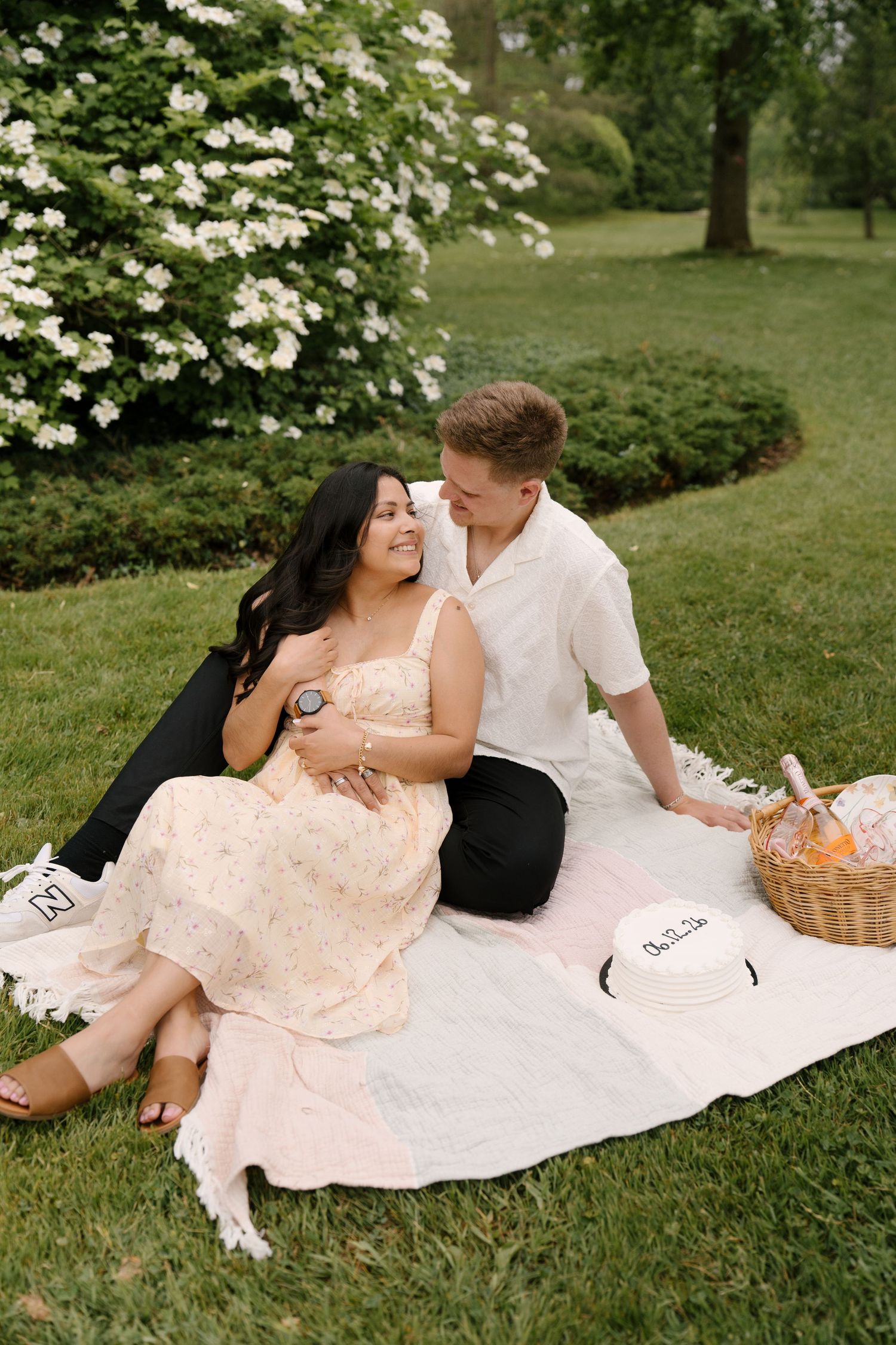 Smiling couple relaxing on a blanket with a picnic basket near white flowering garden shrubs.