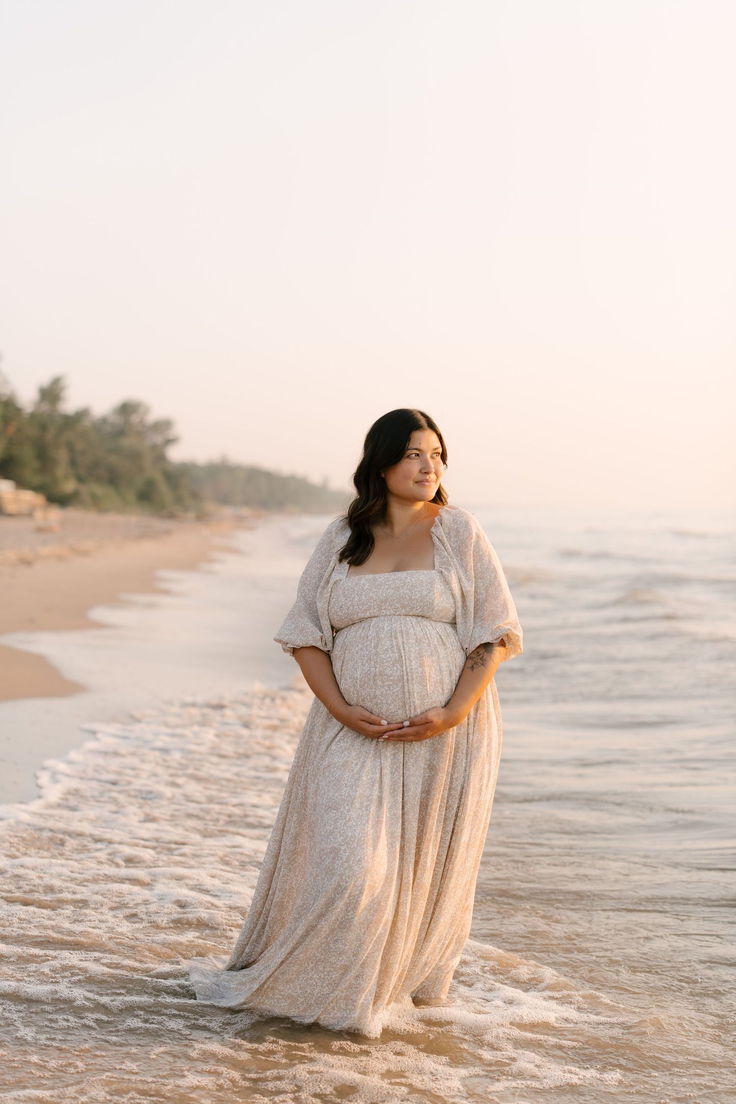 Pregnant woman in flowing white floral dress standing alone on beach shoreline at sunset.
