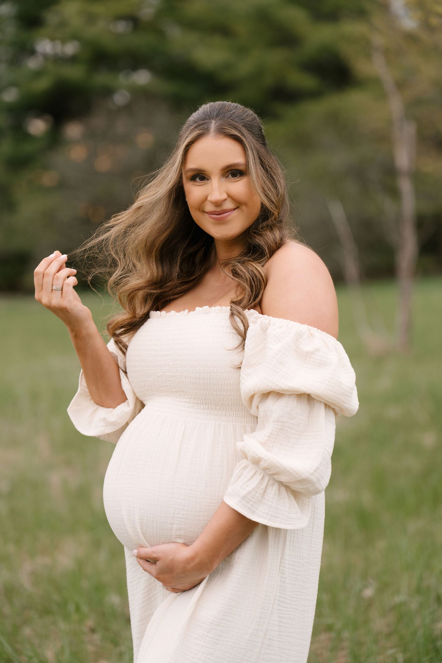 Smiling pregnant woman in white off-shoulder dress holding belly outdoors.