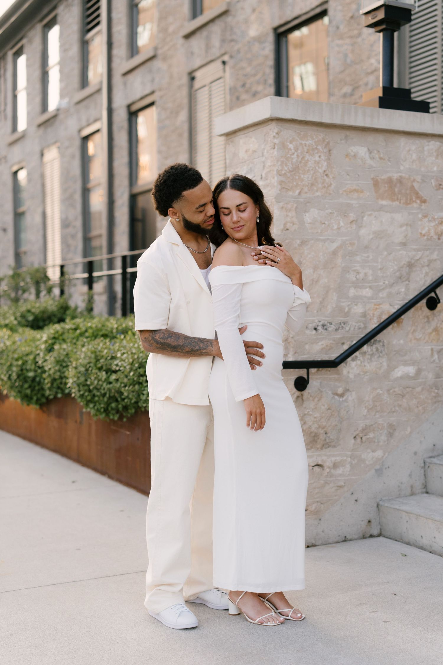 Stylish couple in matching white outfits embracing outside modern stone building at sunset.