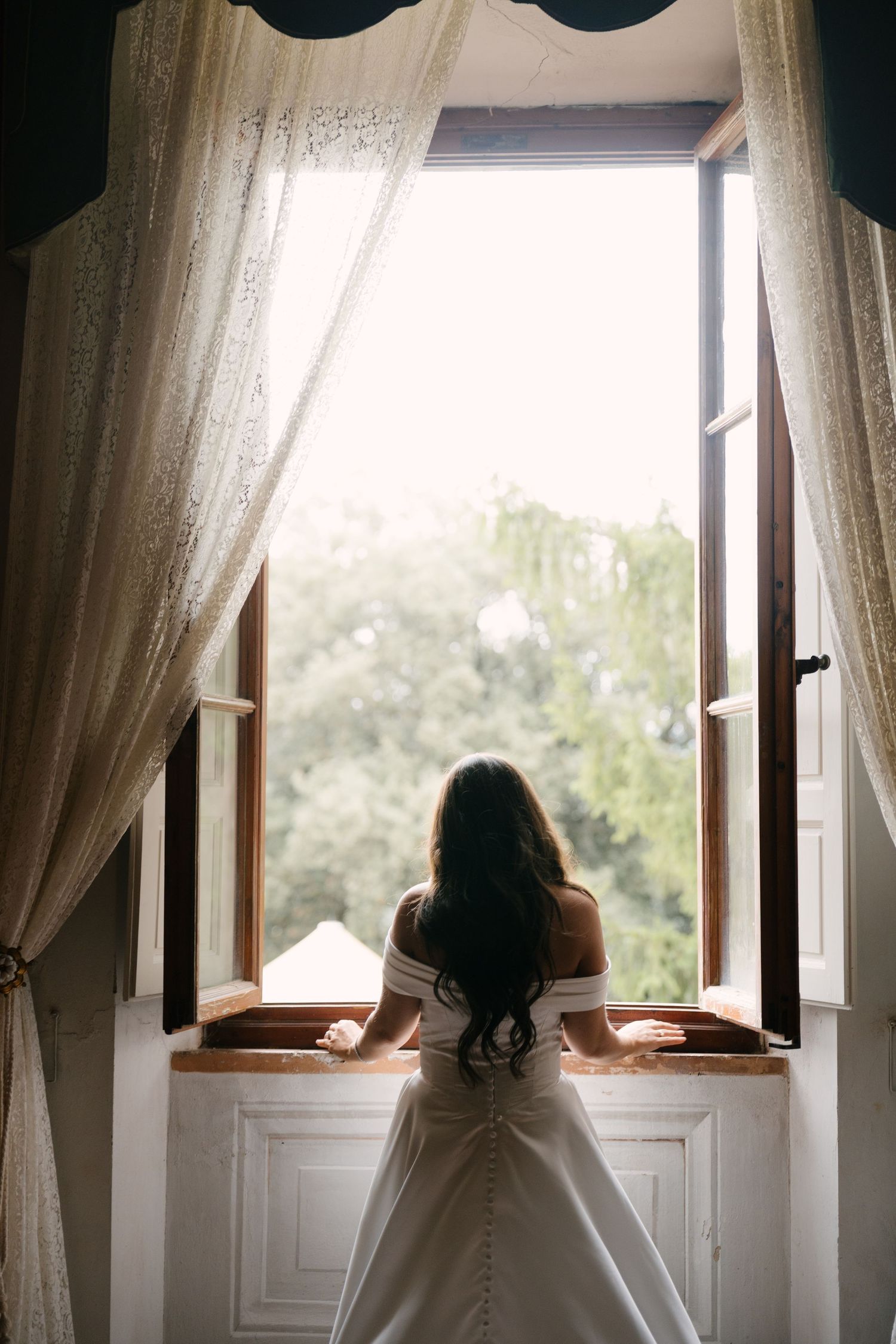 Bride in white off-shoulder gown gazing out an open vintage window with lace curtains and green trees beyond.