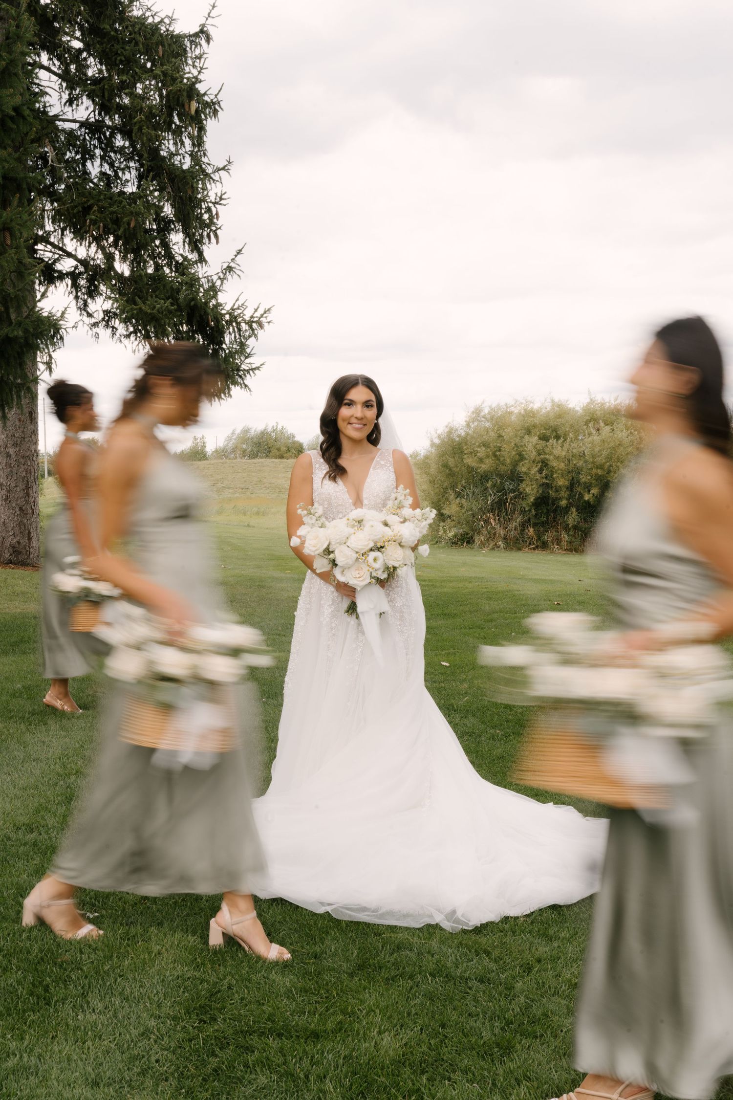 Bride in white ball gown holds white bouquet while bridesmaids in sage dresses walk past in motion blur.