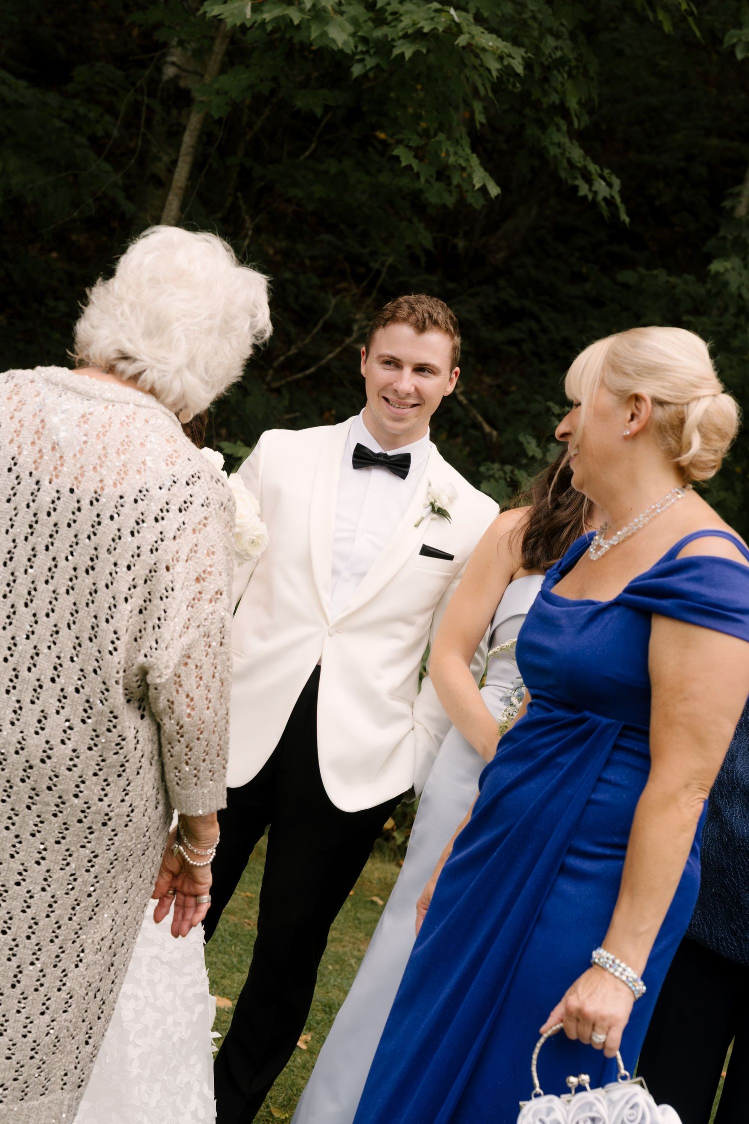 Groom in white tuxedo smiles warmly while greeting elderly and female guests at an outdoor wedding.