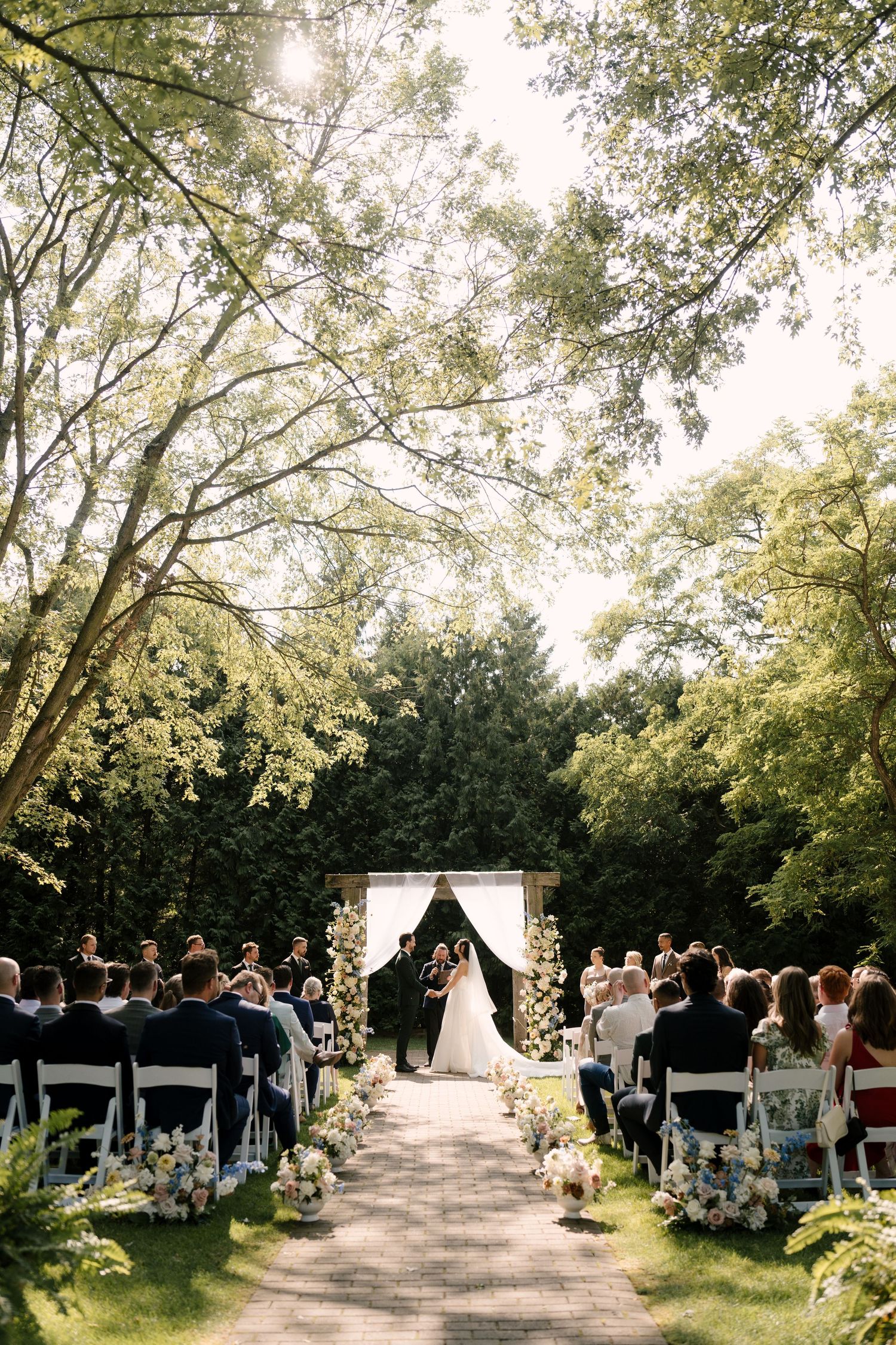 Outdoor garden wedding ceremony with a floral arch, guests seated on white chairs along aisle lined with white flowers at Roseville Estates.