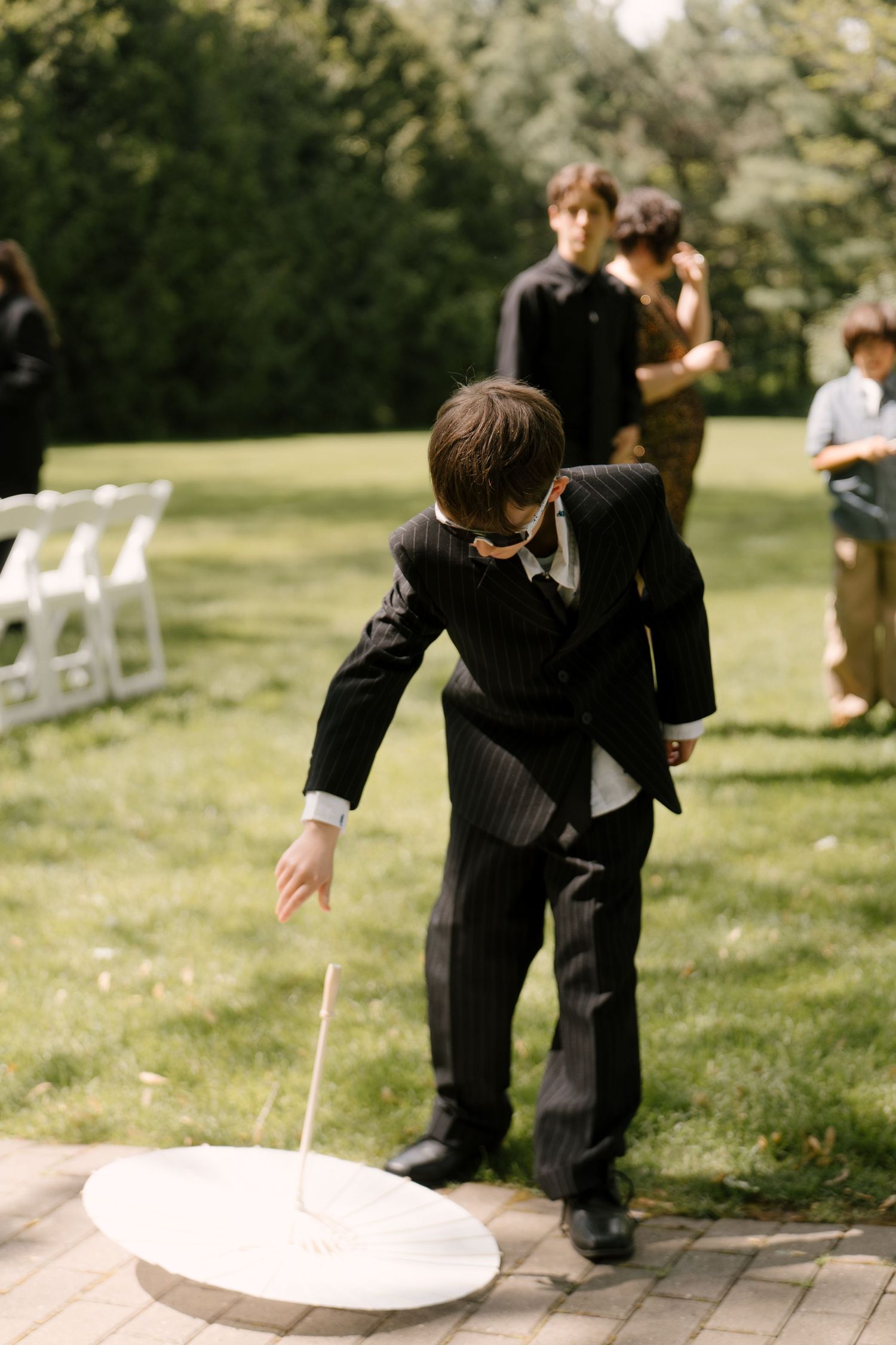 Young ring bearer in black pinstripe suit picks up fallen white parasol on ceremony lawn.
